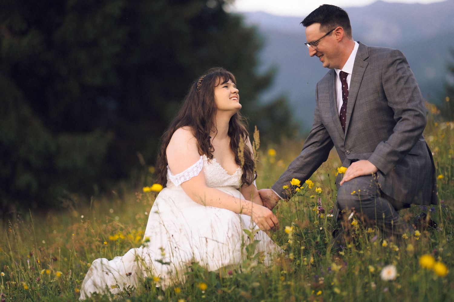 Couple sitting in a wildflower meadow, woman in white dress laughing, man in gray suit, with mountain views.