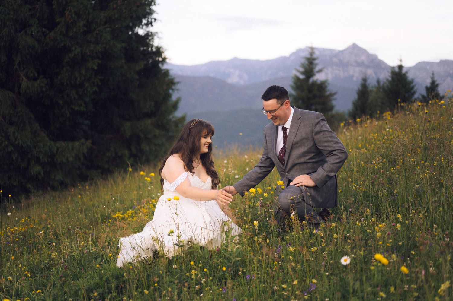 Couple in wedding attire sitting in alpine wildflower meadow with mountain peaks in background.