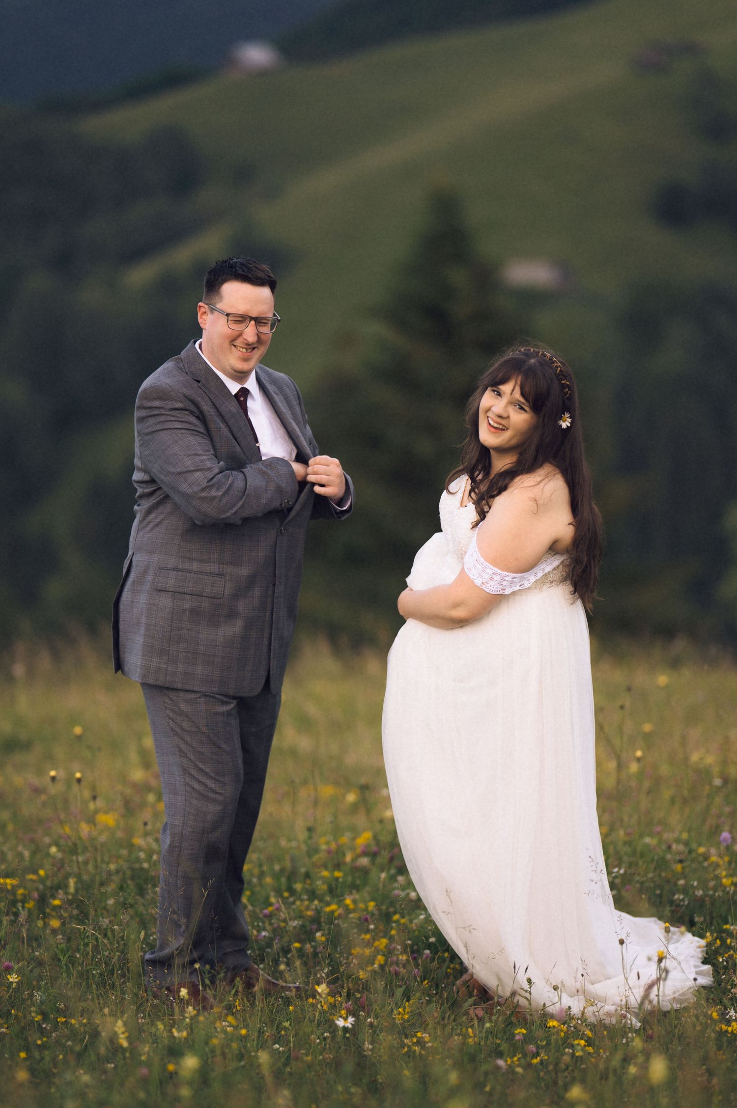 Pregnant bride in white gown stands with groom in gray suit on a green meadow with mountain backdrop.