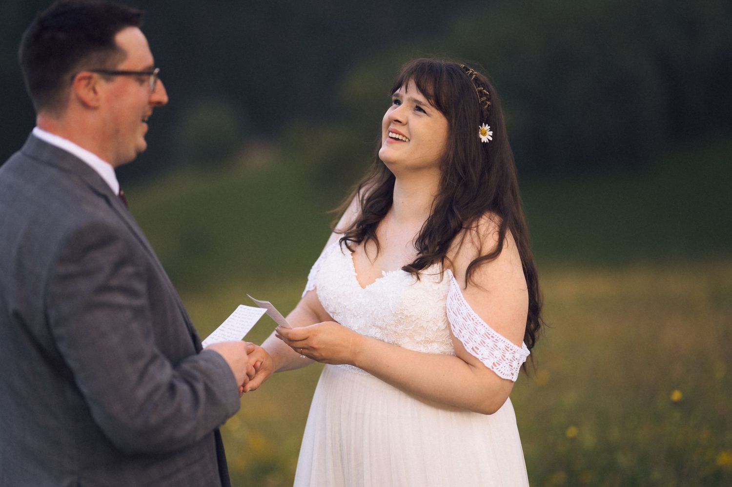 Bride in white off-shoulder gown laughing joyfully during outdoor wedding ceremony vows in a green meadow.