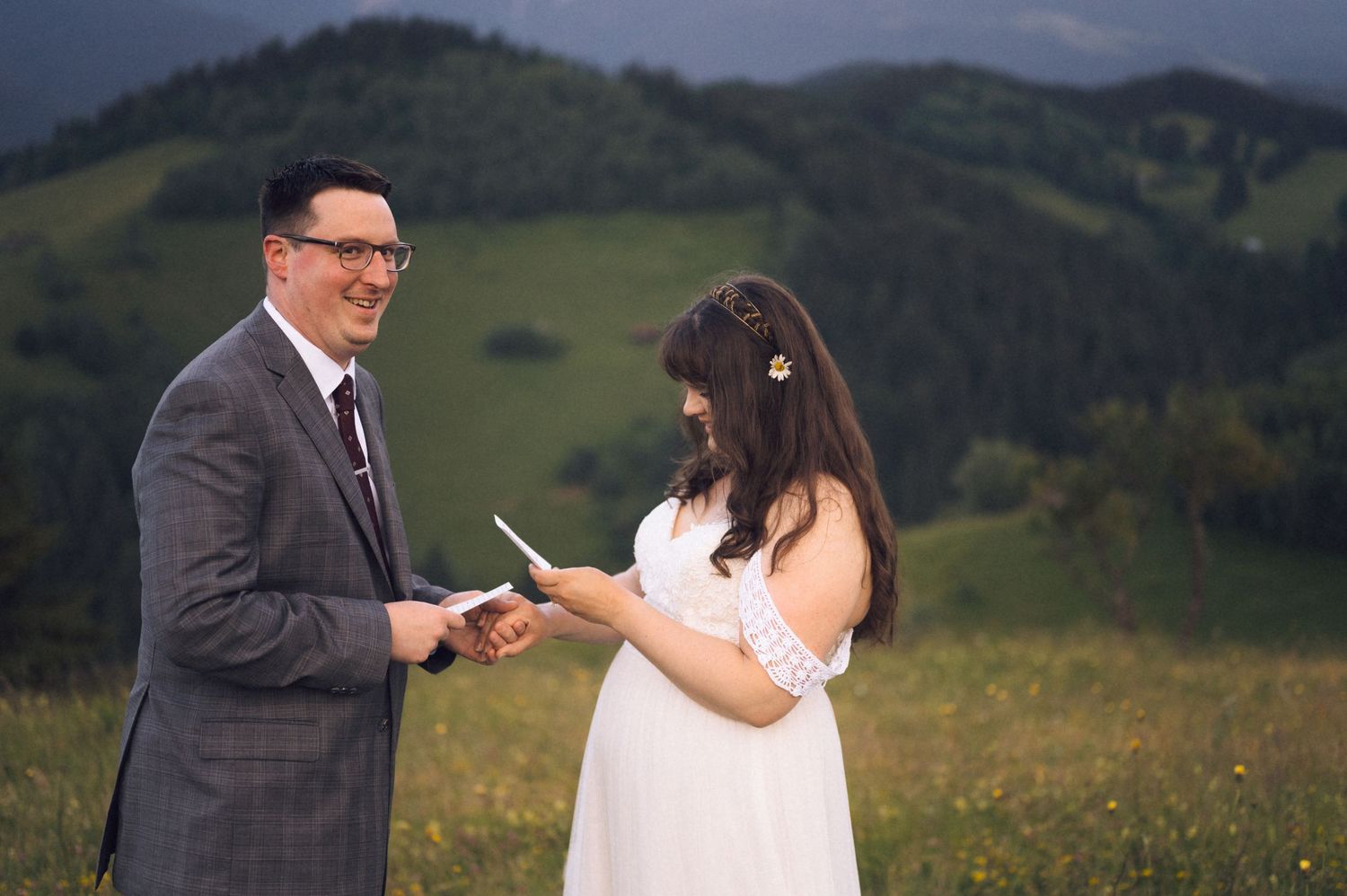 Couple exchanging vows outdoors on a green hillside, bride in white dress reading from paper, groom in gray suit smiling.