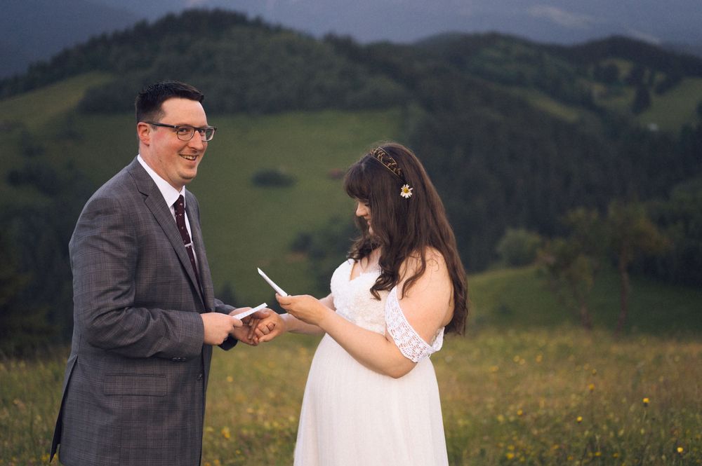 Couple exchanging vows outdoors on a green hillside, bride in white dress reading from paper, groom in gray suit smiling.