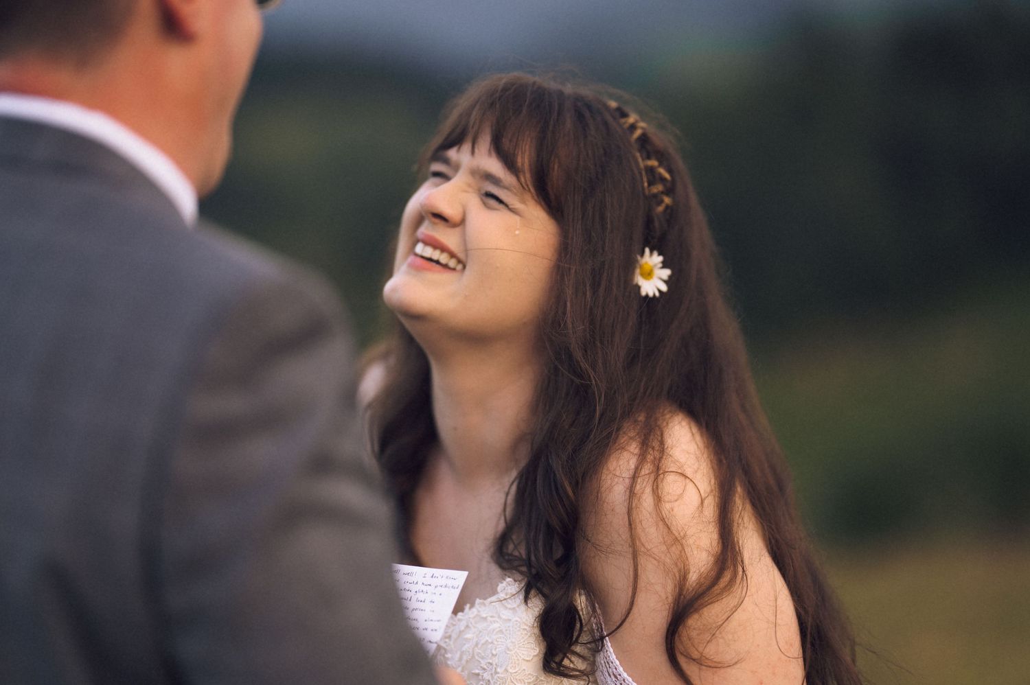 Smiling bride with daisy flower in dark hair laughing joyfully during outdoor wedding ceremony.
