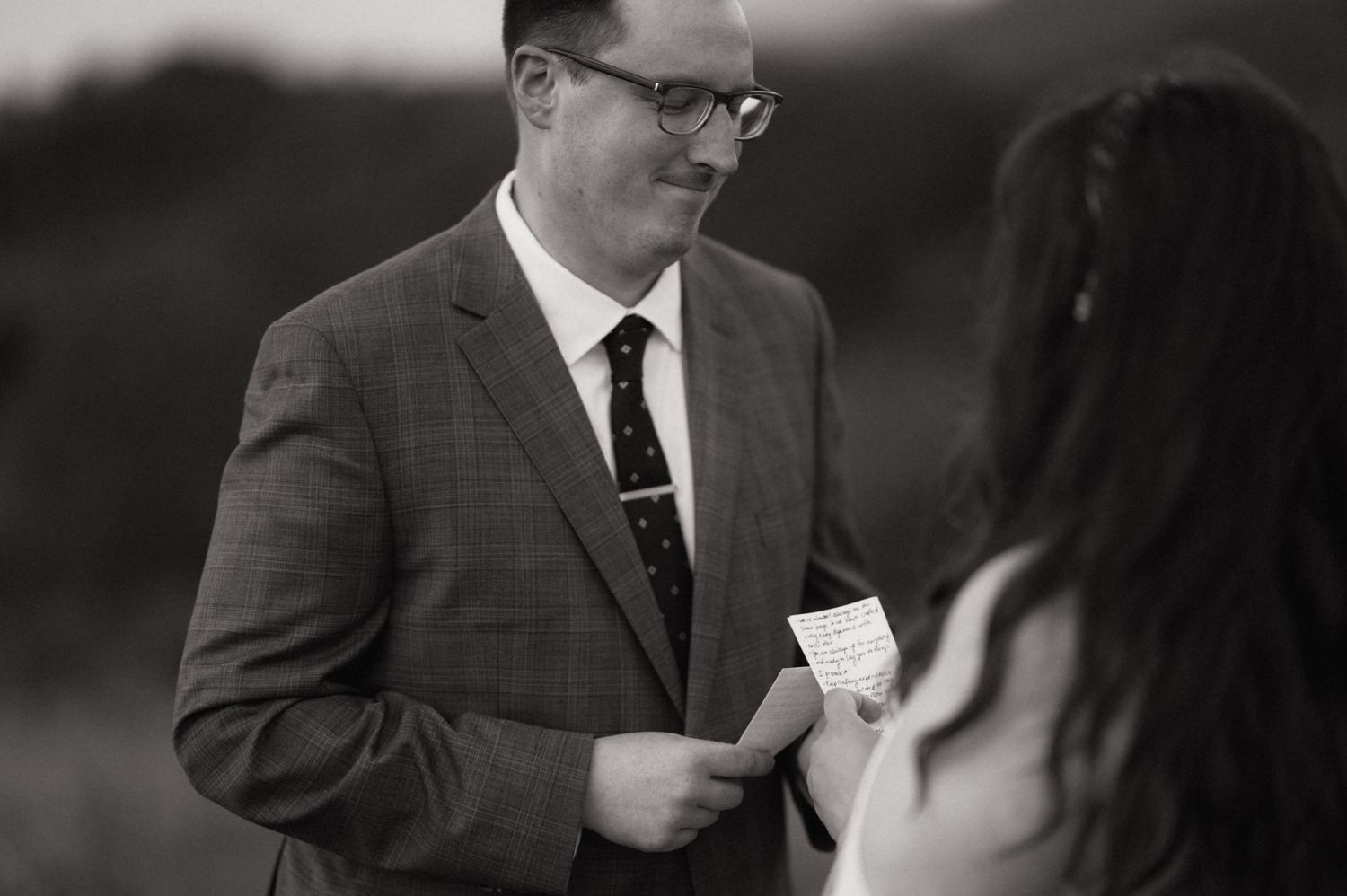 Black and white photo of a groom in a suit reading wedding vows from a card to his bride outdoors.