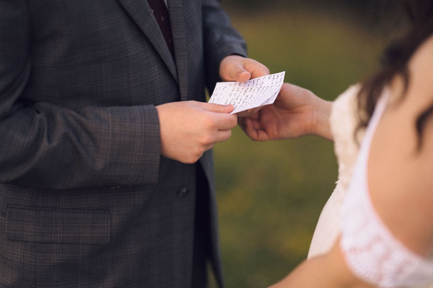 Groom in gray suit exchanging vows card with bride in white dress during outdoor wedding ceremony.