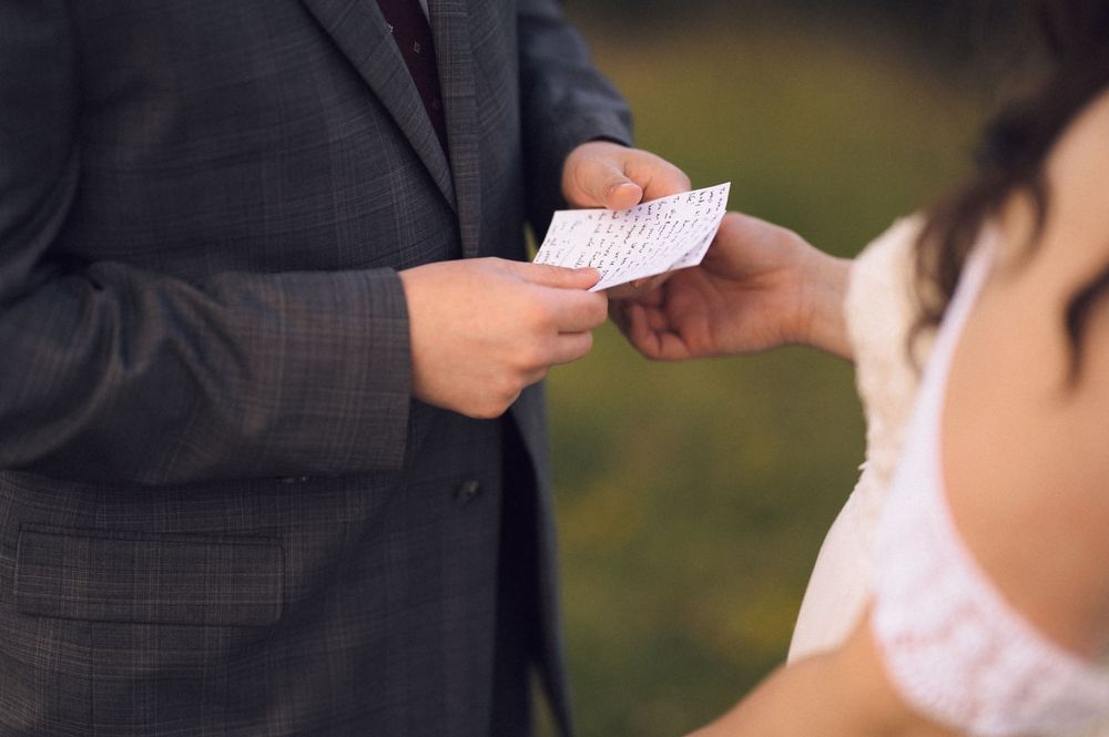 Groom in gray suit exchanging vows card with bride in white dress during outdoor wedding ceremony.