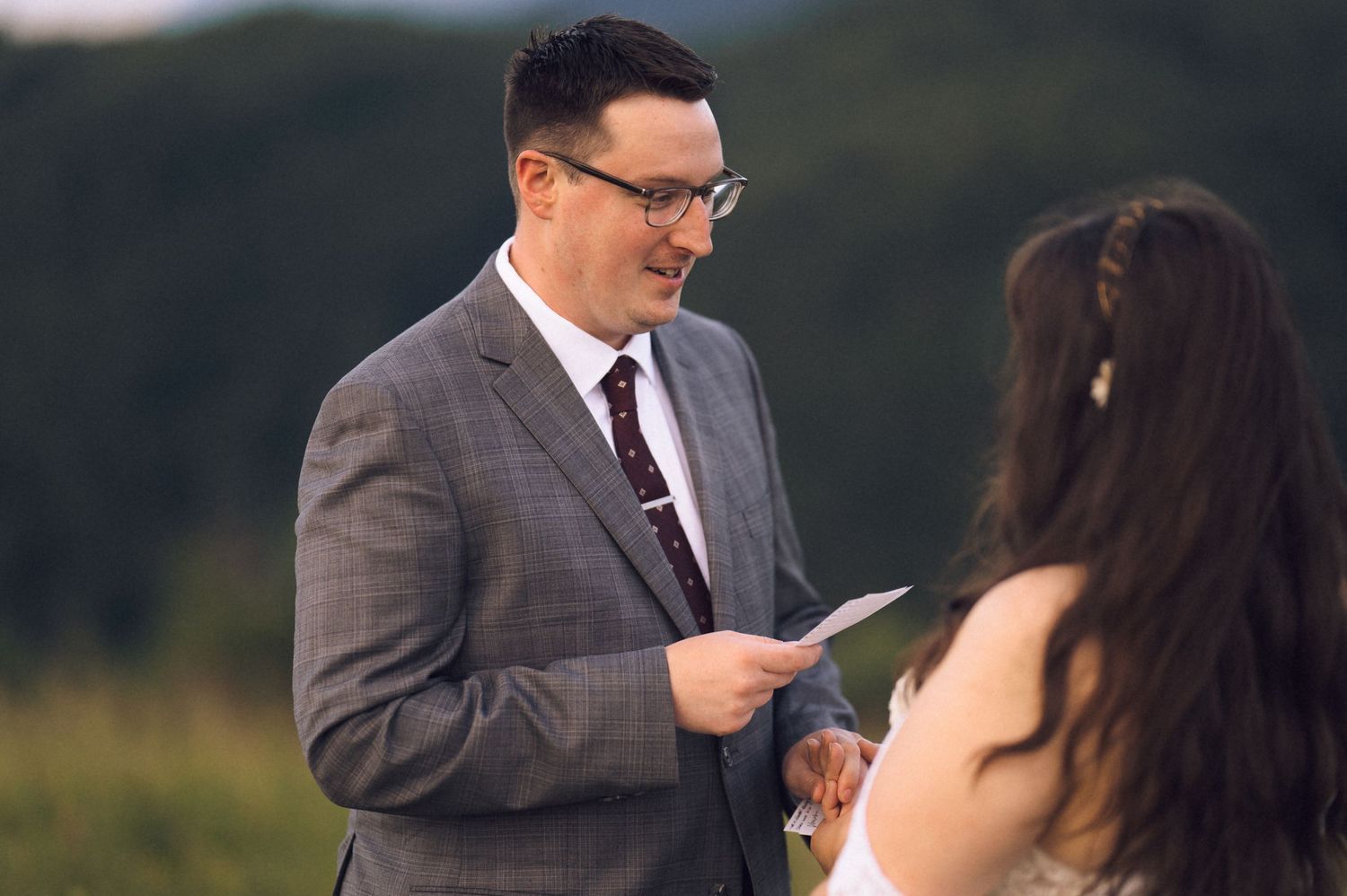 Groom in gray suit reading wedding vows to bride outdoors with green hills in background.