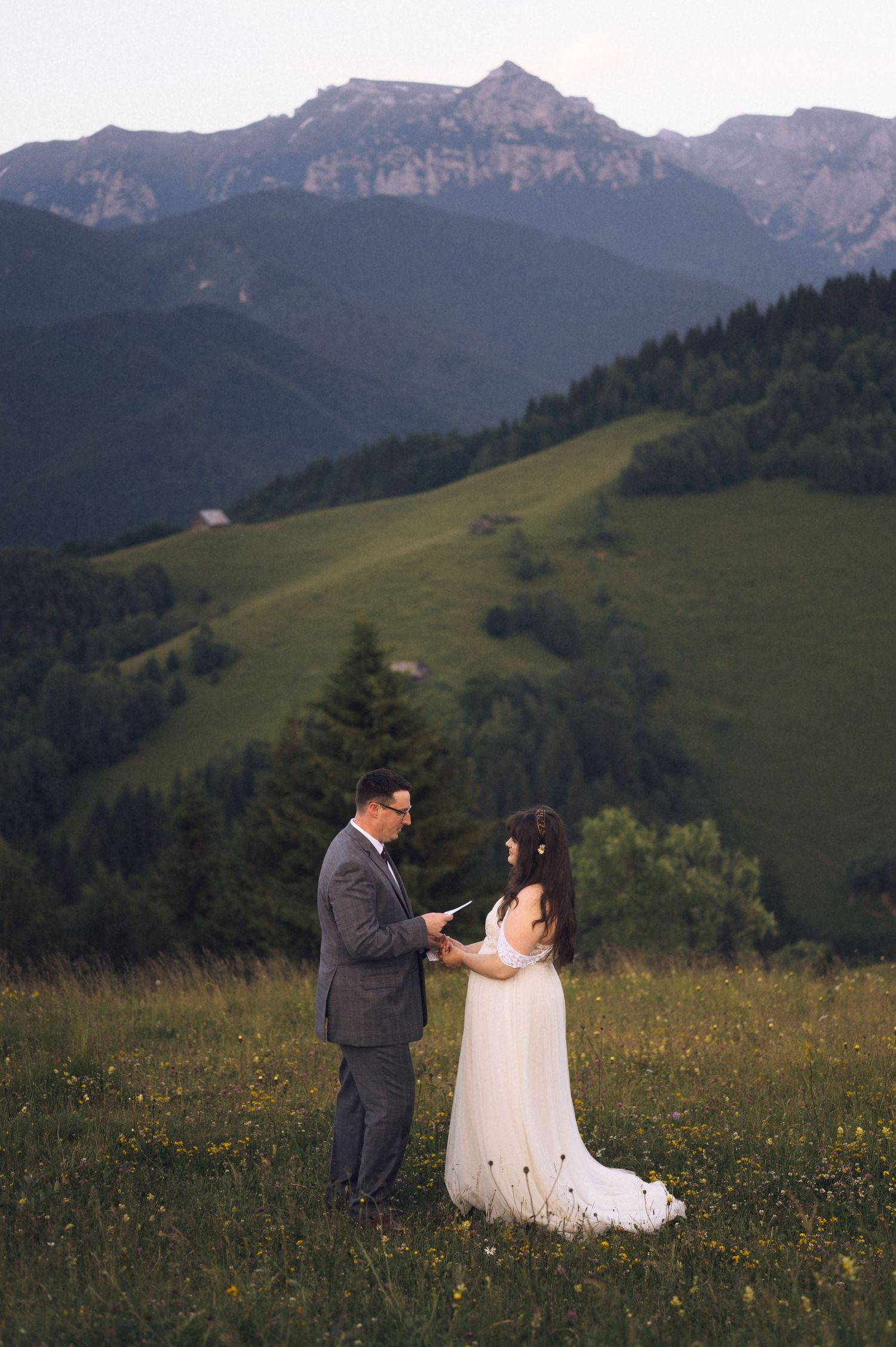 Couple exchanging vows in a mountain meadow, bride in white gown, groom in gray suit, lush green hills backdrop.