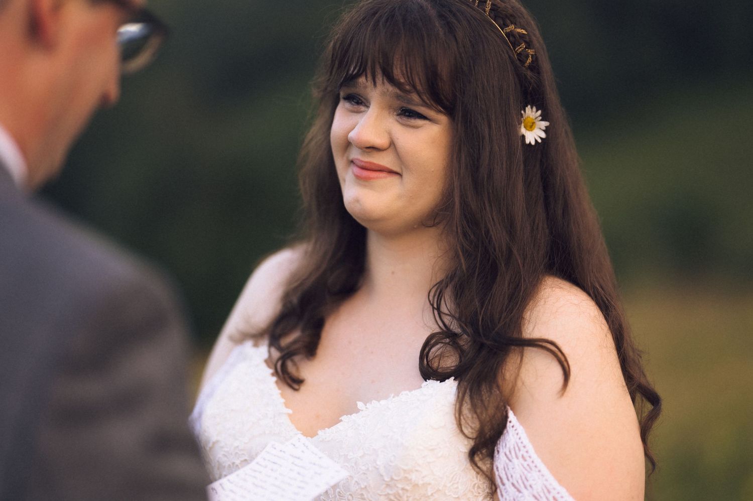 Bride with dark hair and daisy hair accessory smiling warmly during outdoor wedding ceremony.