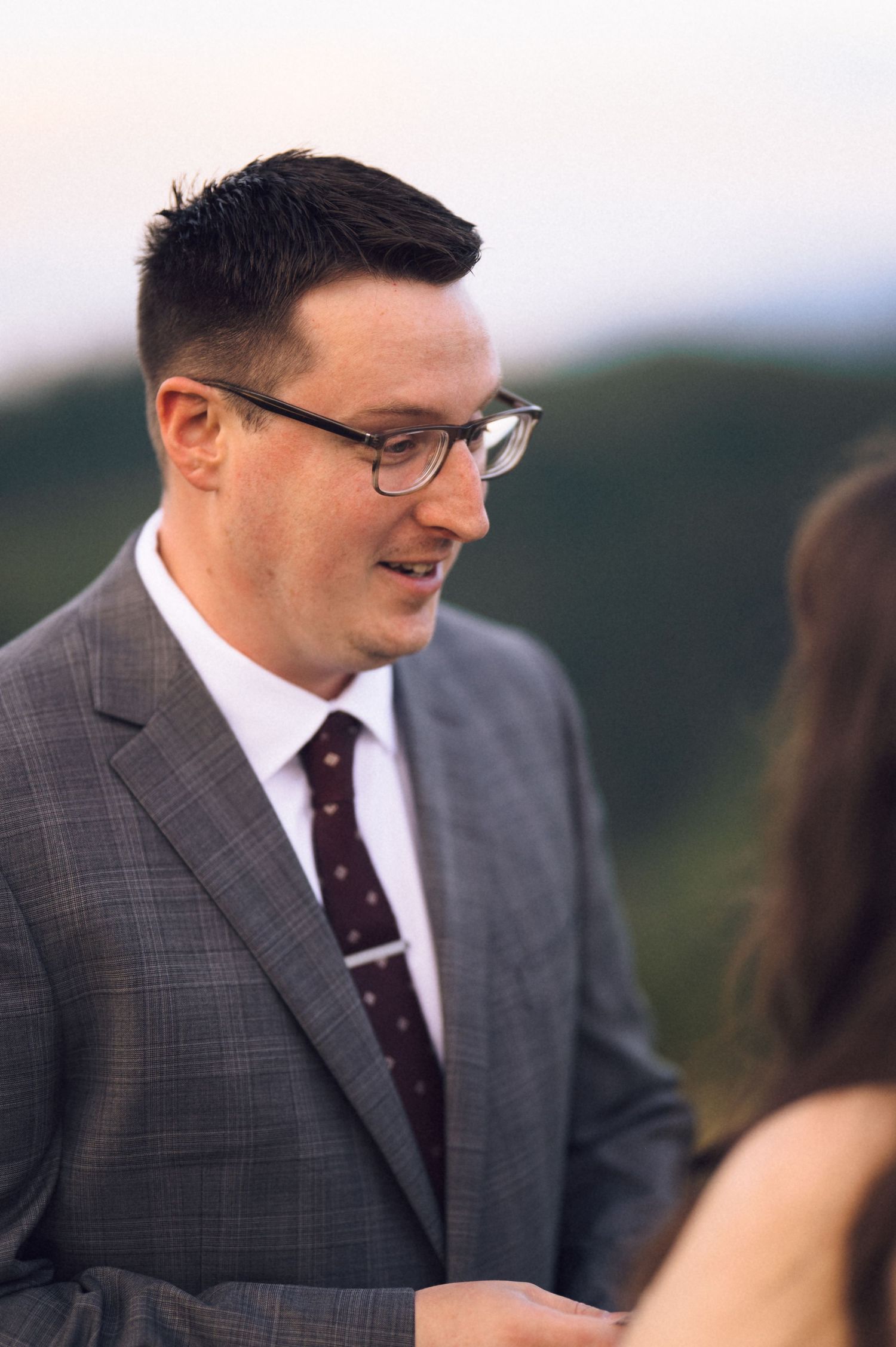 A groom in a gray suit and burgundy tie smiles during an outdoor wedding ceremony with green hills behind him.