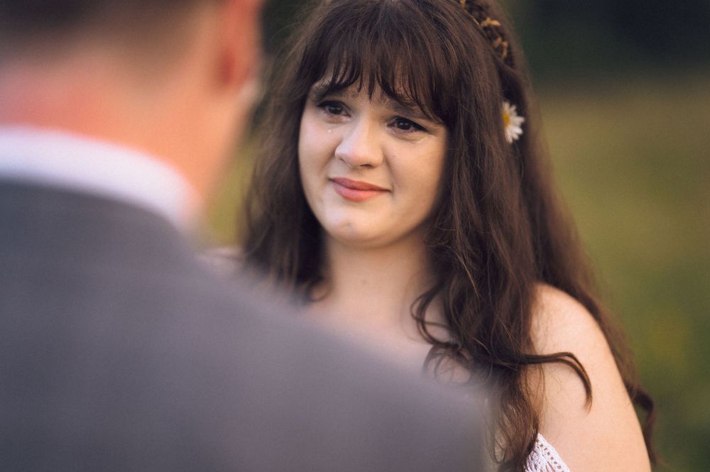 A young woman with dark hair and a daisy flower crown smiles warmly during an outdoor wedding ceremony.