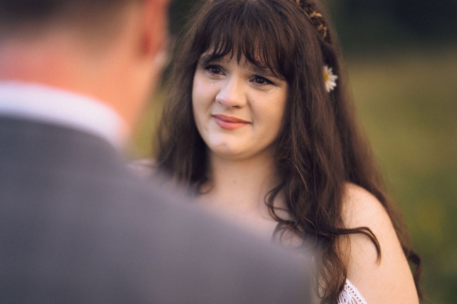 A young woman with dark hair and a daisy flower crown smiles warmly during an outdoor wedding ceremony.