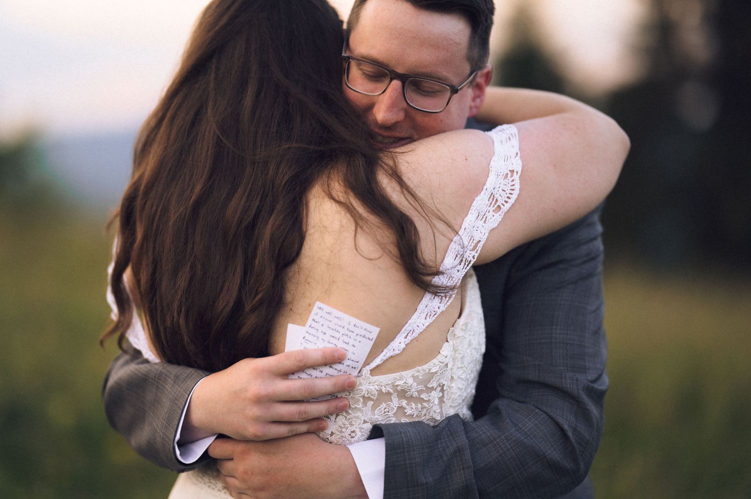 Bride and groom share an emotional hug outdoors, man holding note card, woman in lace wedding dress.