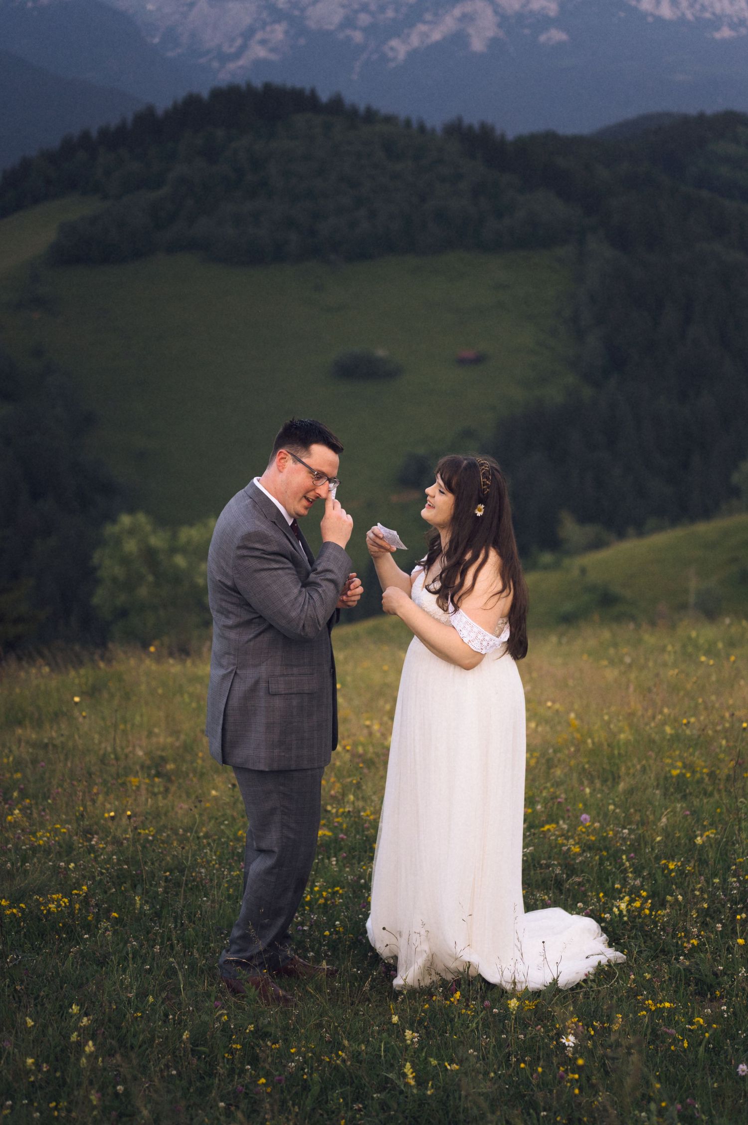 Bride and groom share emotional moment in alpine meadow, groom wiping tears in gray suit, bride in white gown.