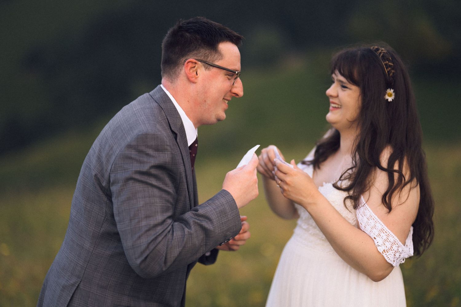 Couple exchanging vows outdoors in a lush green field, bride in white dress and groom in gray suit smiling.