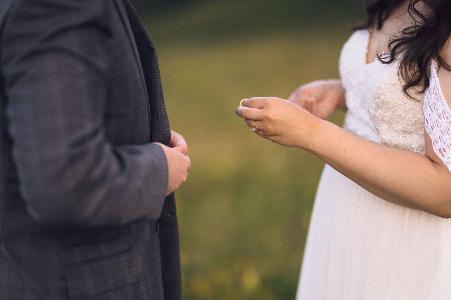 Bride holding a wedding ring during an outdoor ring exchange ceremony with groom in gray suit.