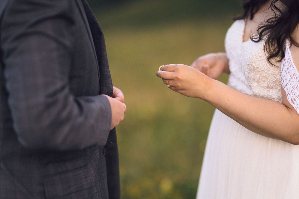 Bride holding a wedding ring during an outdoor ring exchange ceremony with groom in gray suit.