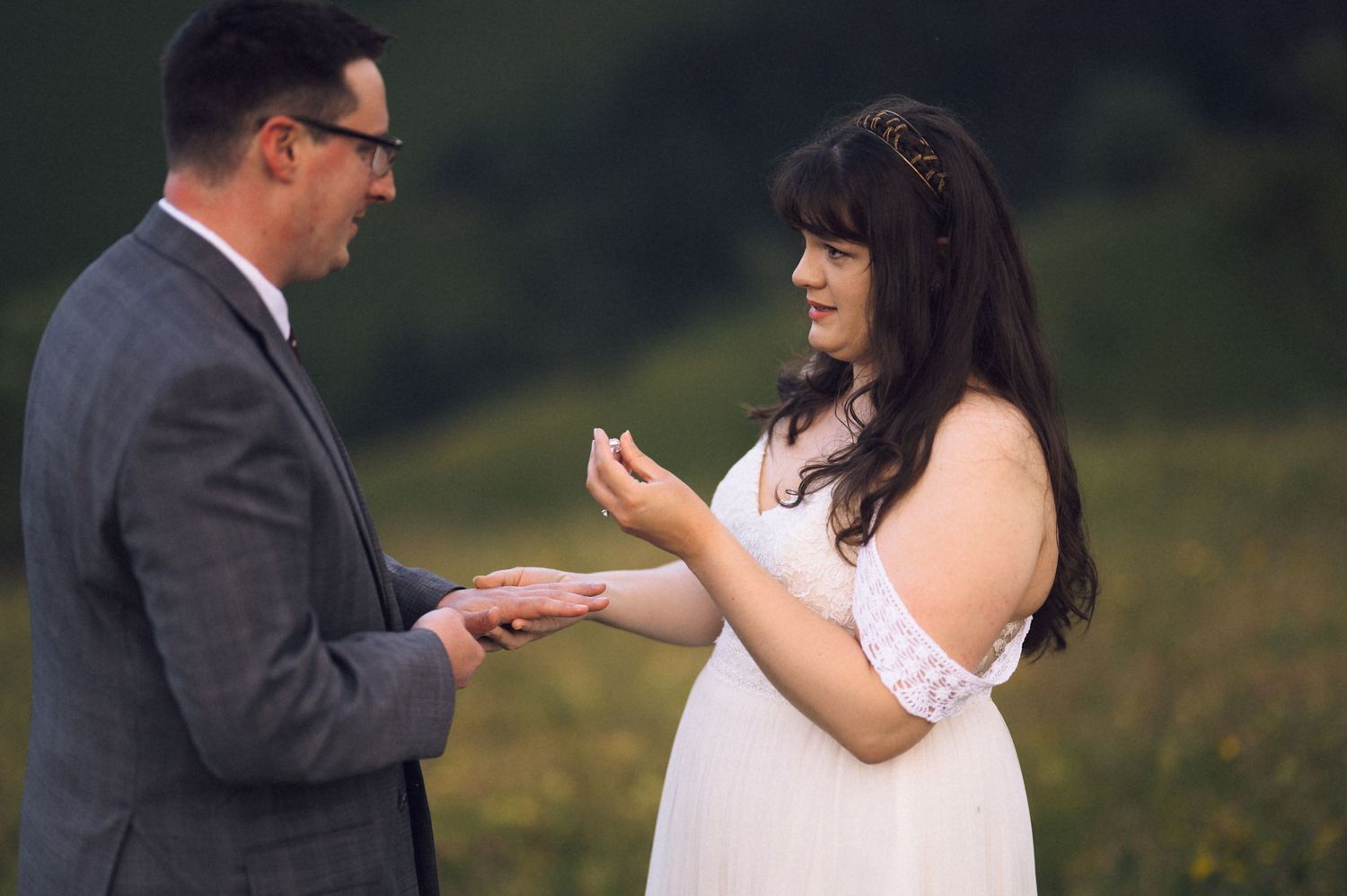 Bride placing wedding ring on groom's finger during outdoor ceremony in green meadow.