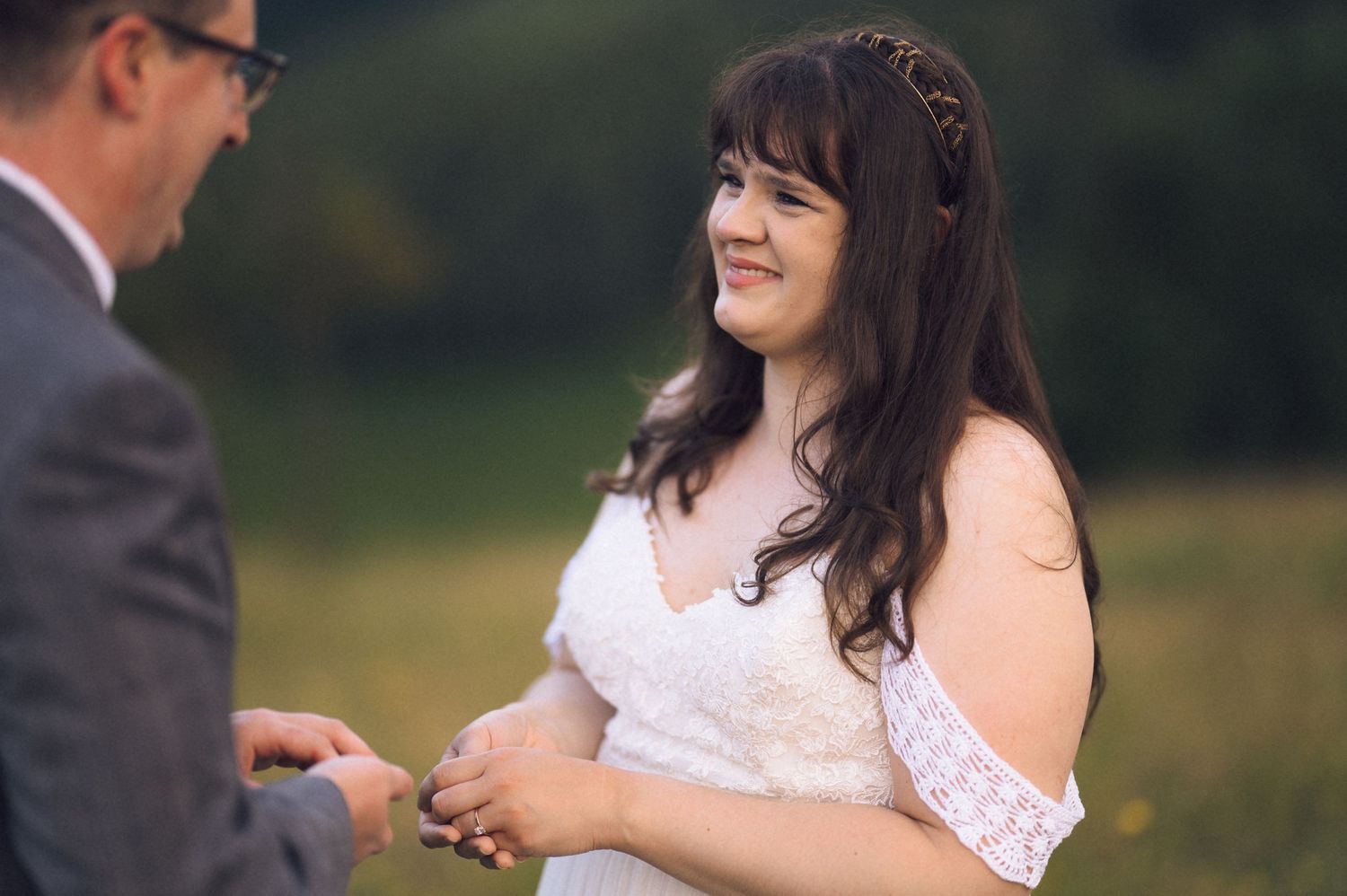 Bride in white lace dress smiling during outdoor wedding ceremony in a green field.