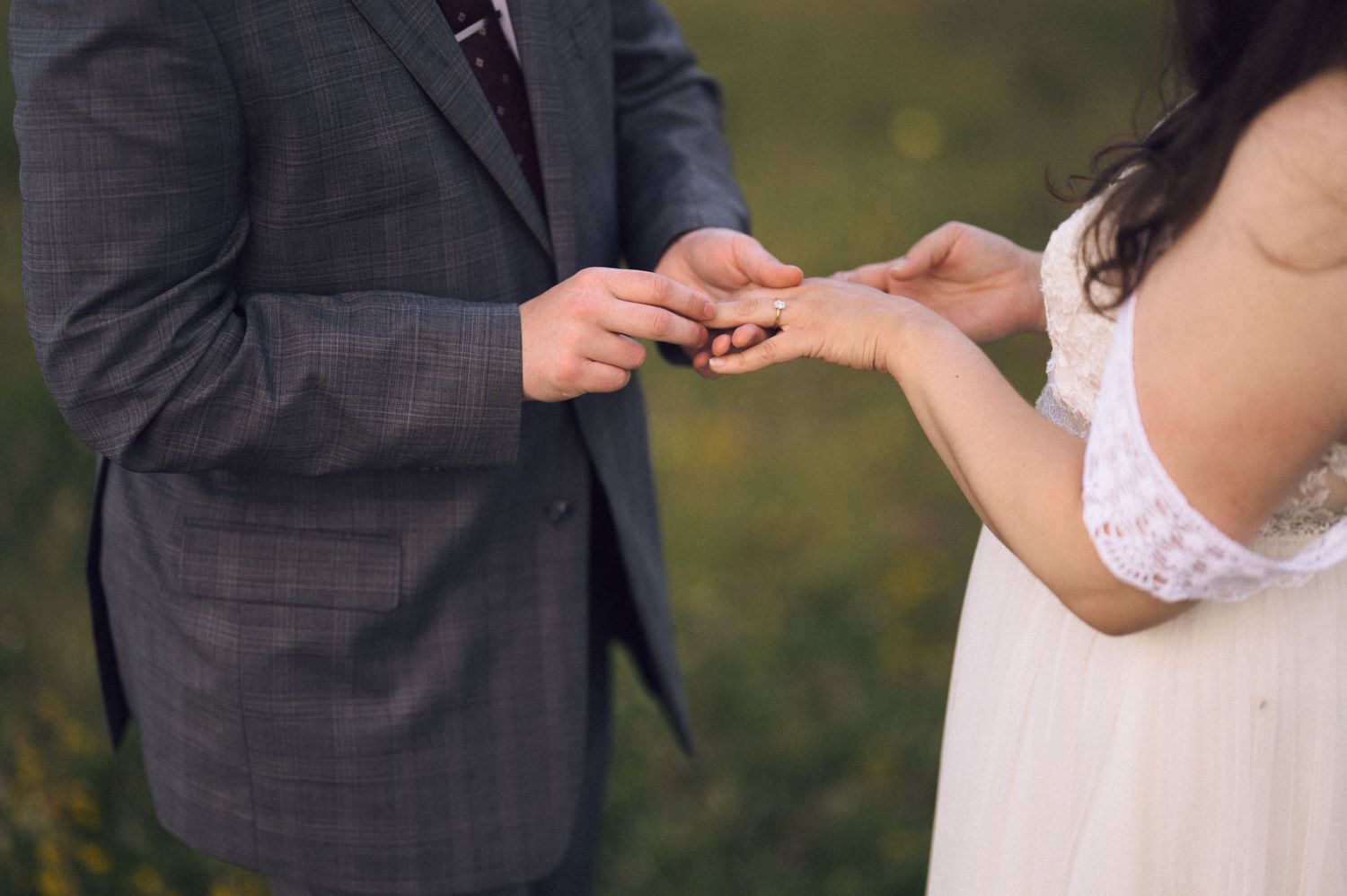 Bride placing a wedding ring on groom's finger during outdoor ceremony, couple in elegant attire exchanging vows.