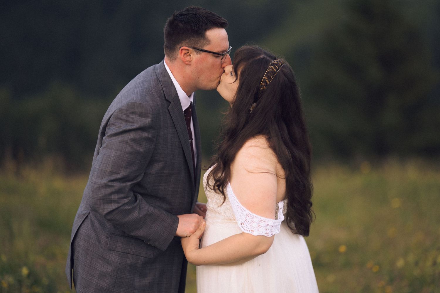 Couple sharing a romantic kiss in a lush green meadow, groom in gray suit and bride in white off-shoulder gown.