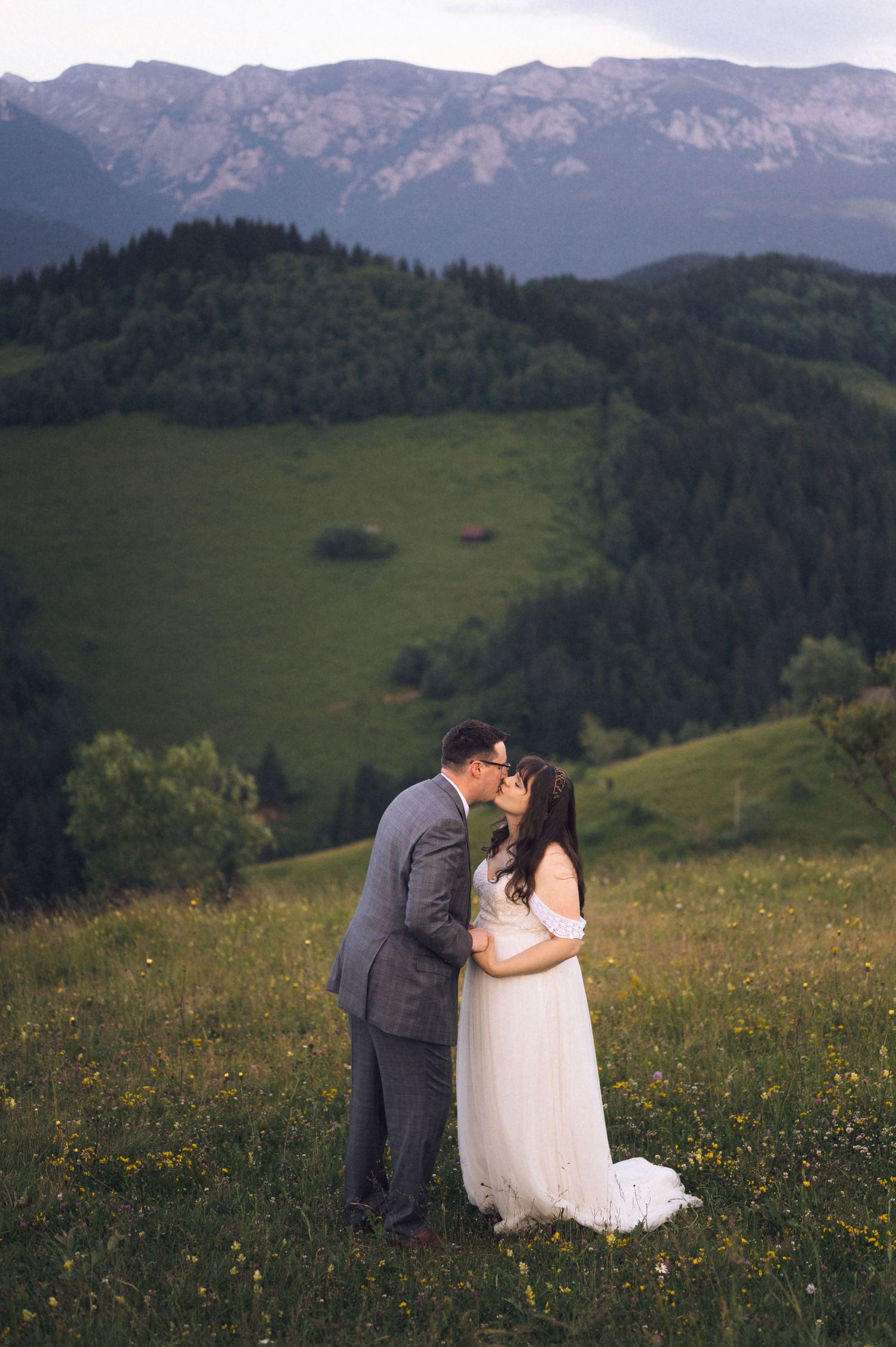 Bride and groom kissing in a green meadow with rolling hills and mountains in the background.