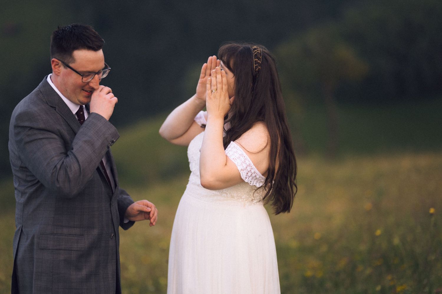 Emotional bride covering her face during a wedding first look with groom in a sunlit meadow.