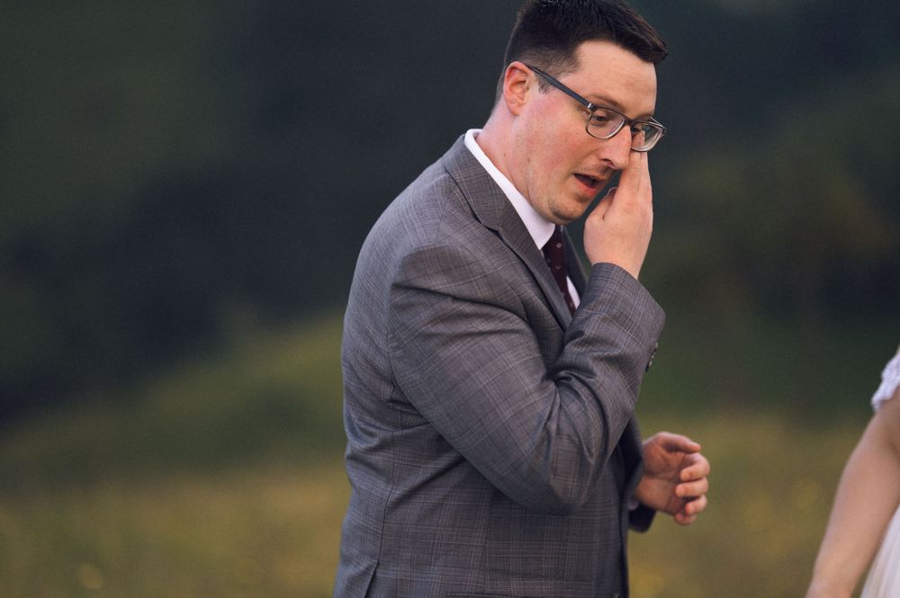 Emotional groom in gray suit wiping tears at outdoor wedding ceremony with green hills in background.