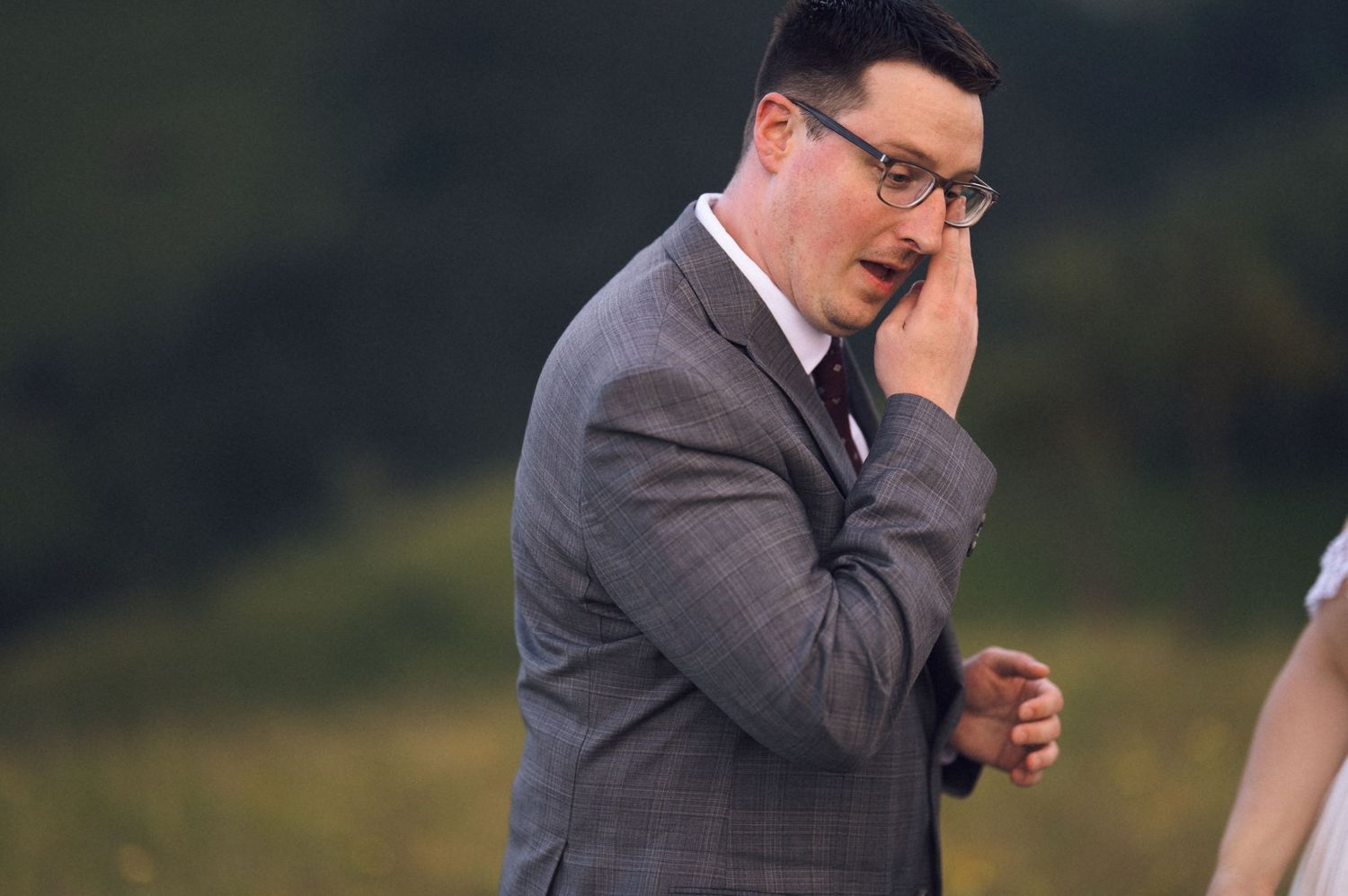 Emotional groom in gray suit wiping tears at outdoor wedding ceremony with green hills in background.