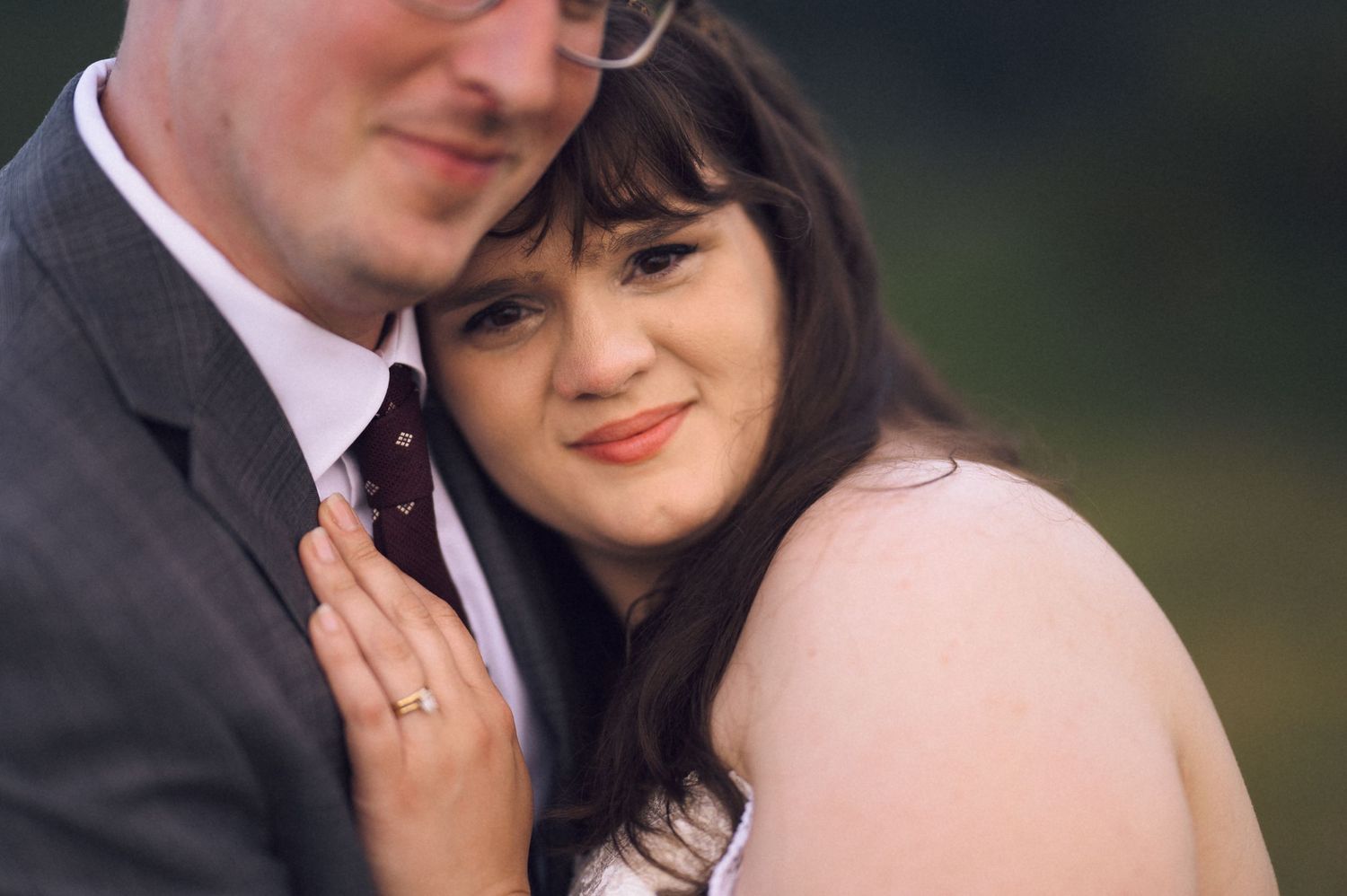 A couple in formal wedding attire embraces closely, the woman wearing a ring and leaning against the man in a gray suit.