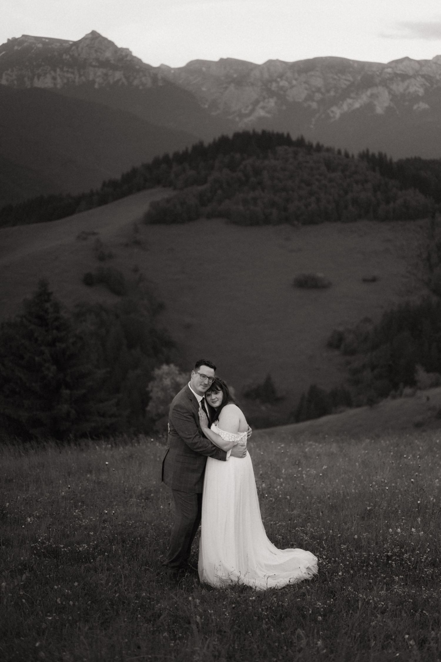 Black and white wedding photo of couple embracing in alpine meadow with mountain peaks and forest in background.