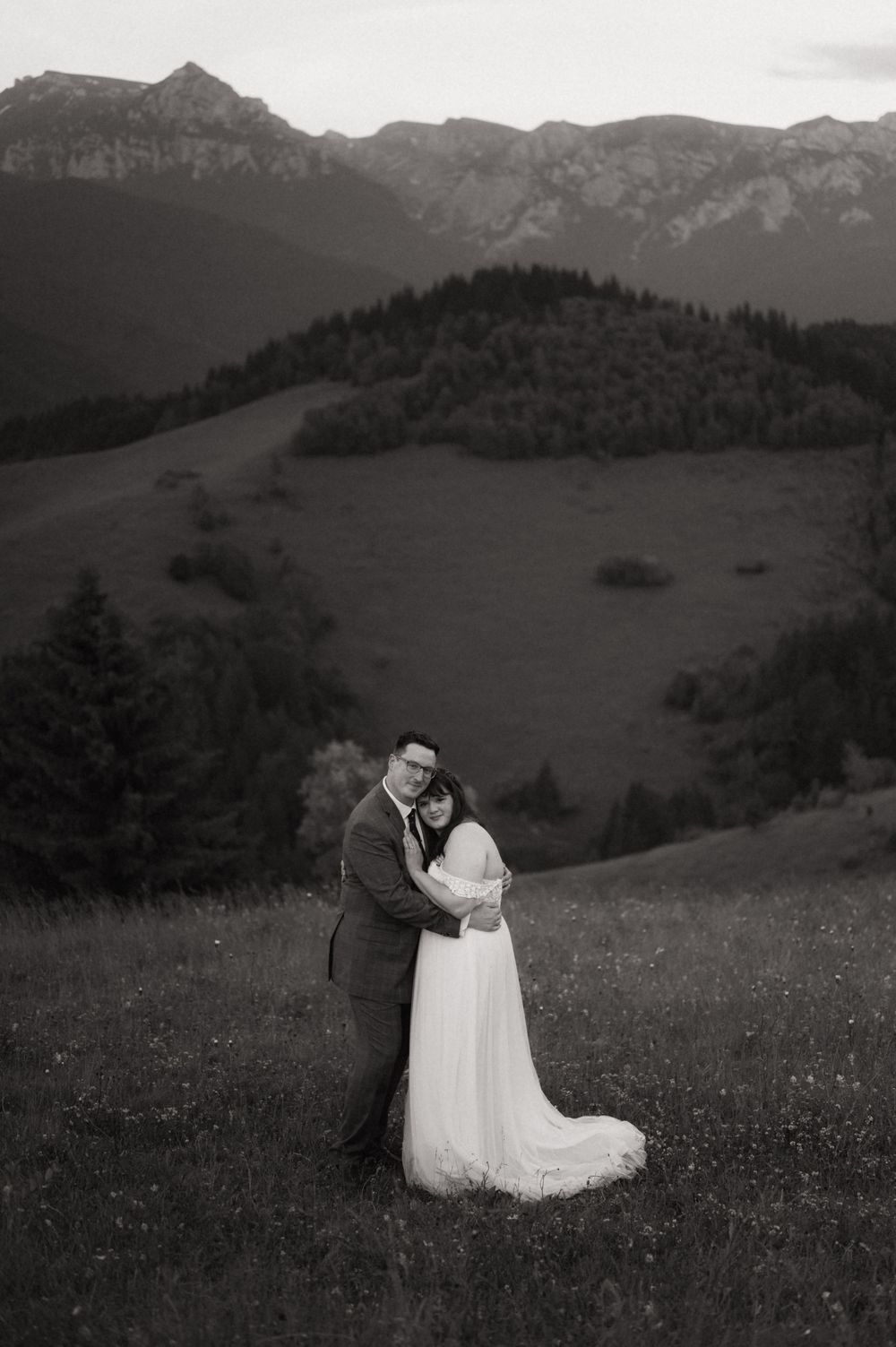 Black and white wedding photo of couple embracing in alpine meadow with mountain peaks and forest in background.