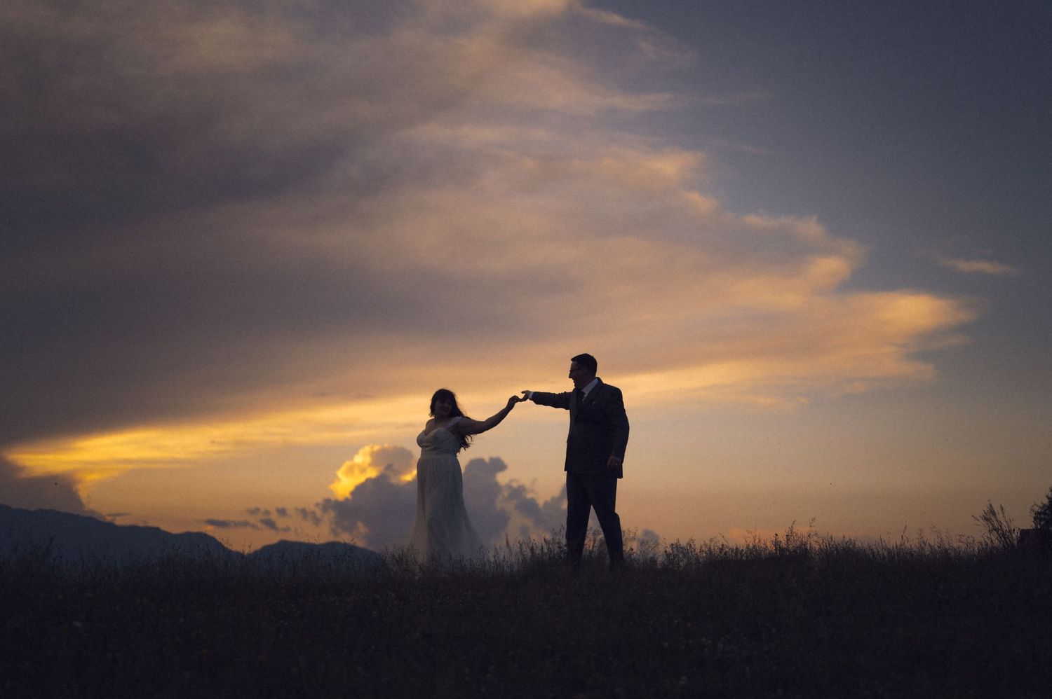 Silhouette of a couple holding hands at sunset with dramatic clouds and mountains in the background.