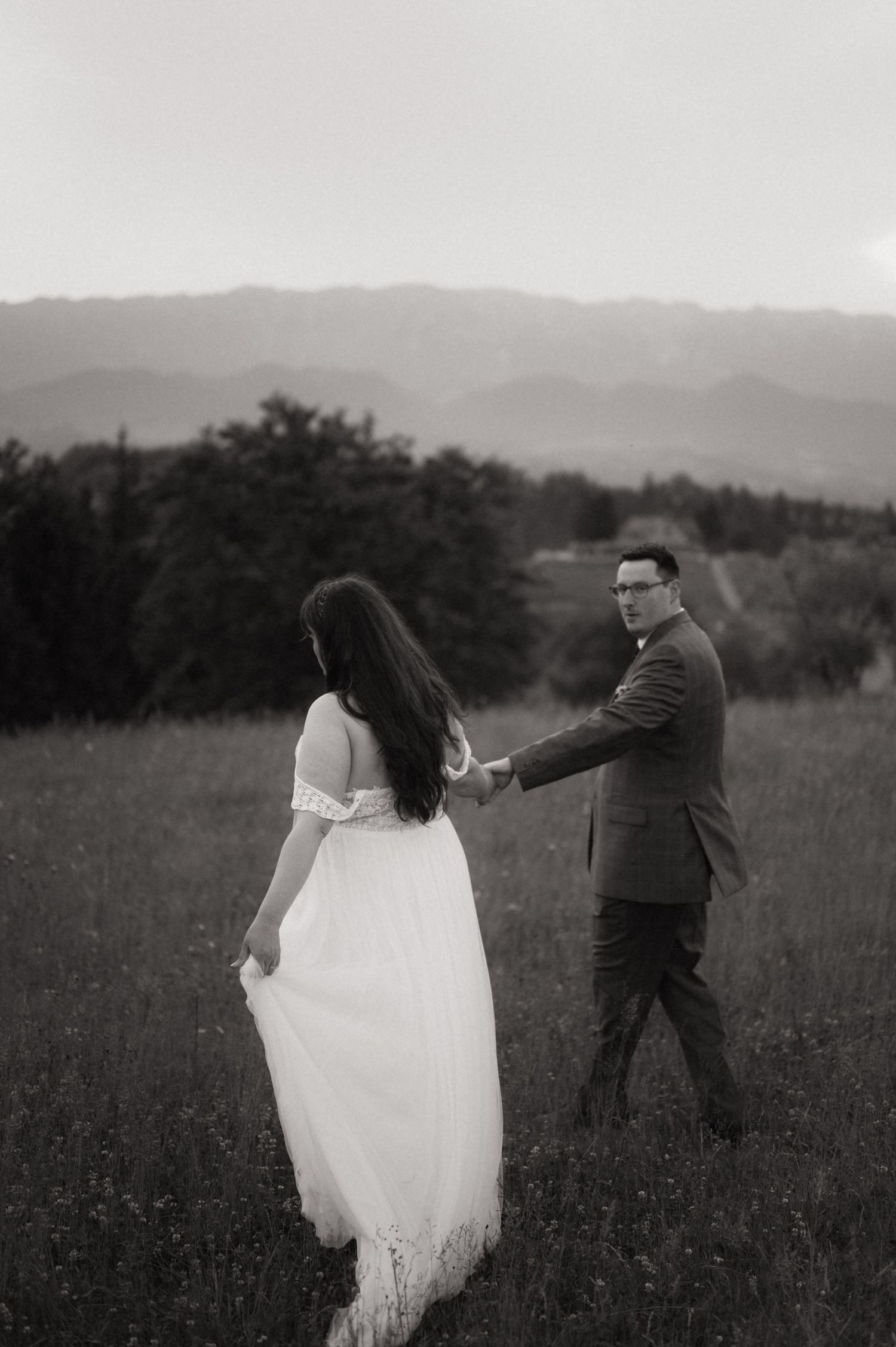 Black and white photo of a couple holding hands in a meadow with mountains in the background.