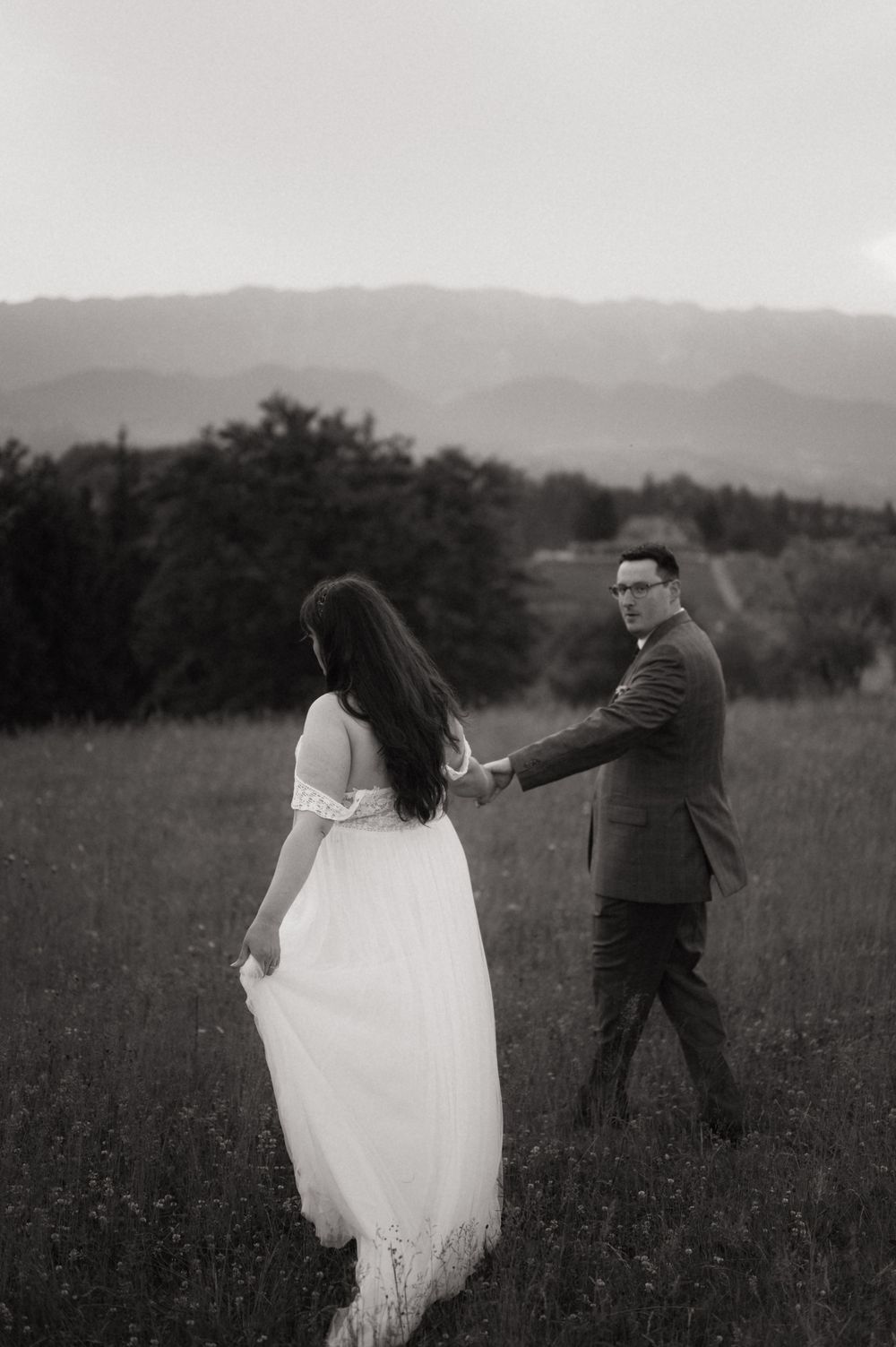 Black and white photo of a couple holding hands in a meadow with mountains in the background.