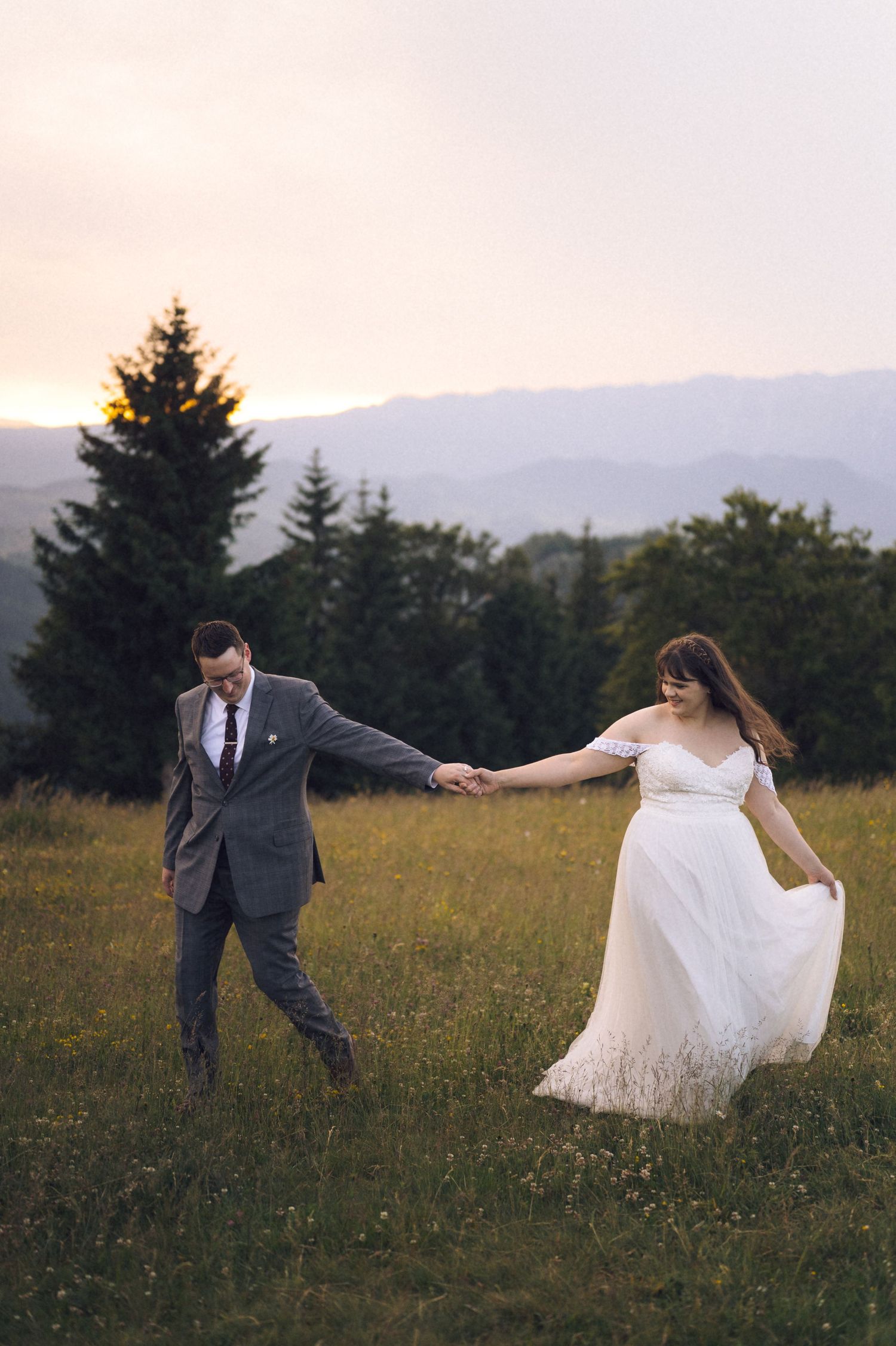 Bride and groom holding hands dancing in a meadow at sunset with mountain views and evergreen trees.