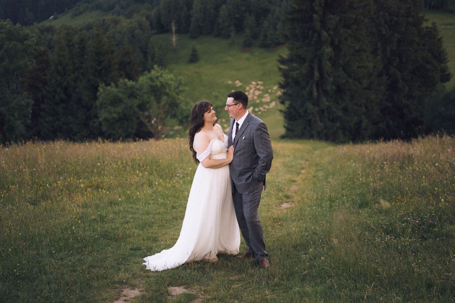 A bride and groom share a romantic moment in a lush green meadow surrounded by pine trees on their wedding day.