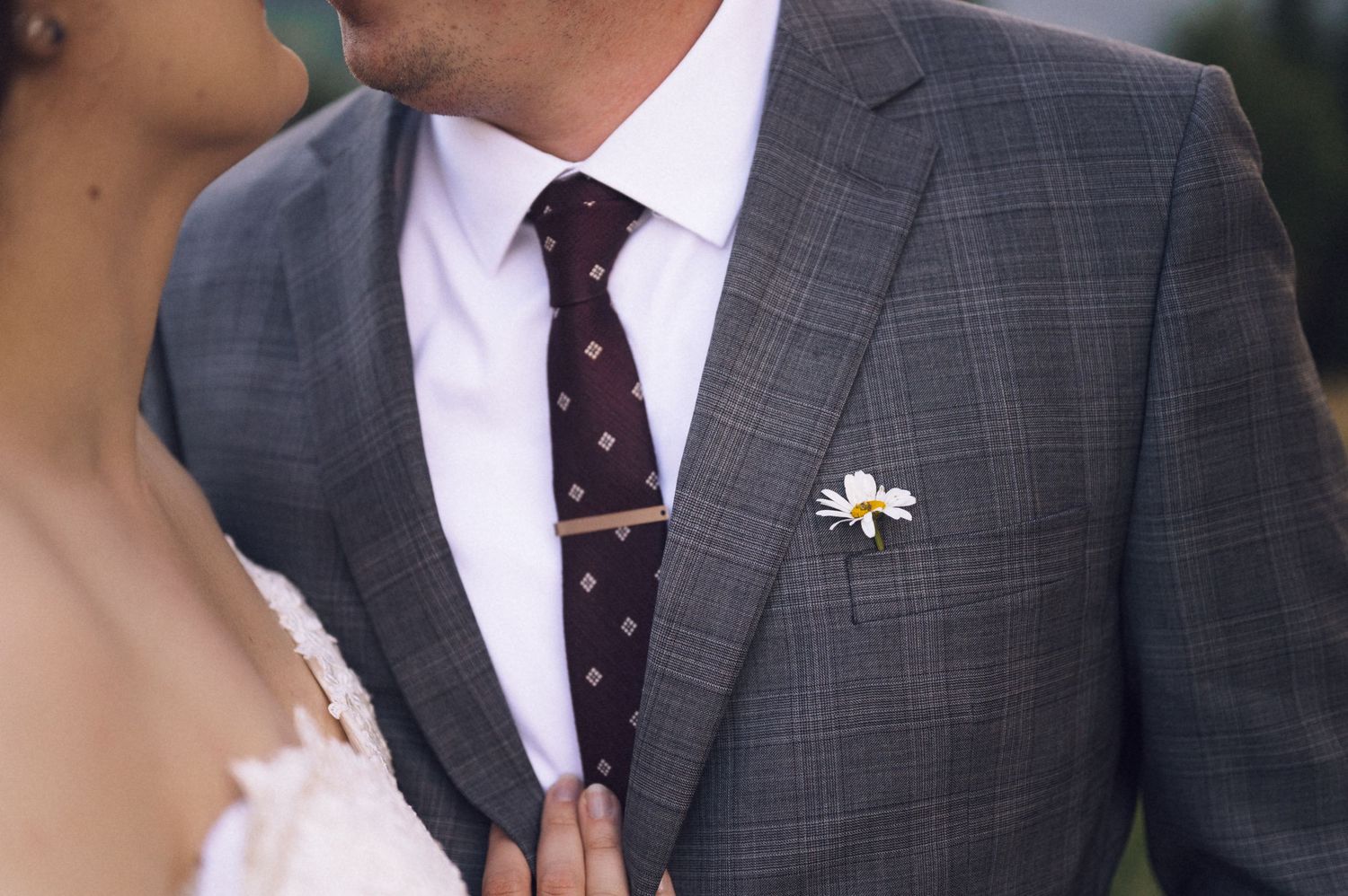Groom in gray plaid suit with burgundy dotted tie and daisy boutonniere, standing beside bride in white dress.