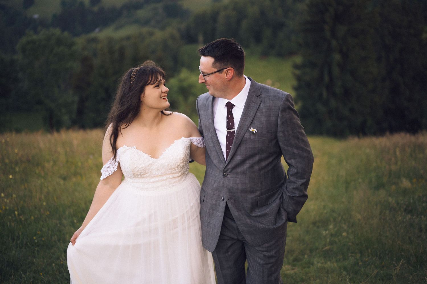 Bride in white off-shoulder gown and groom in gray suit walk through green meadow, sharing a loving glance.