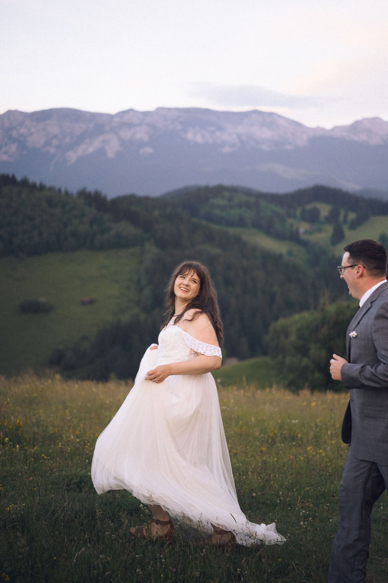 Pregnant bride in white gown smiles at groom on scenic mountain hillside.