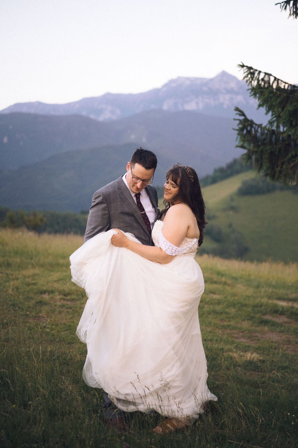Groom lifts bride in white off-shoulder gown on mountain meadow at dusk.
