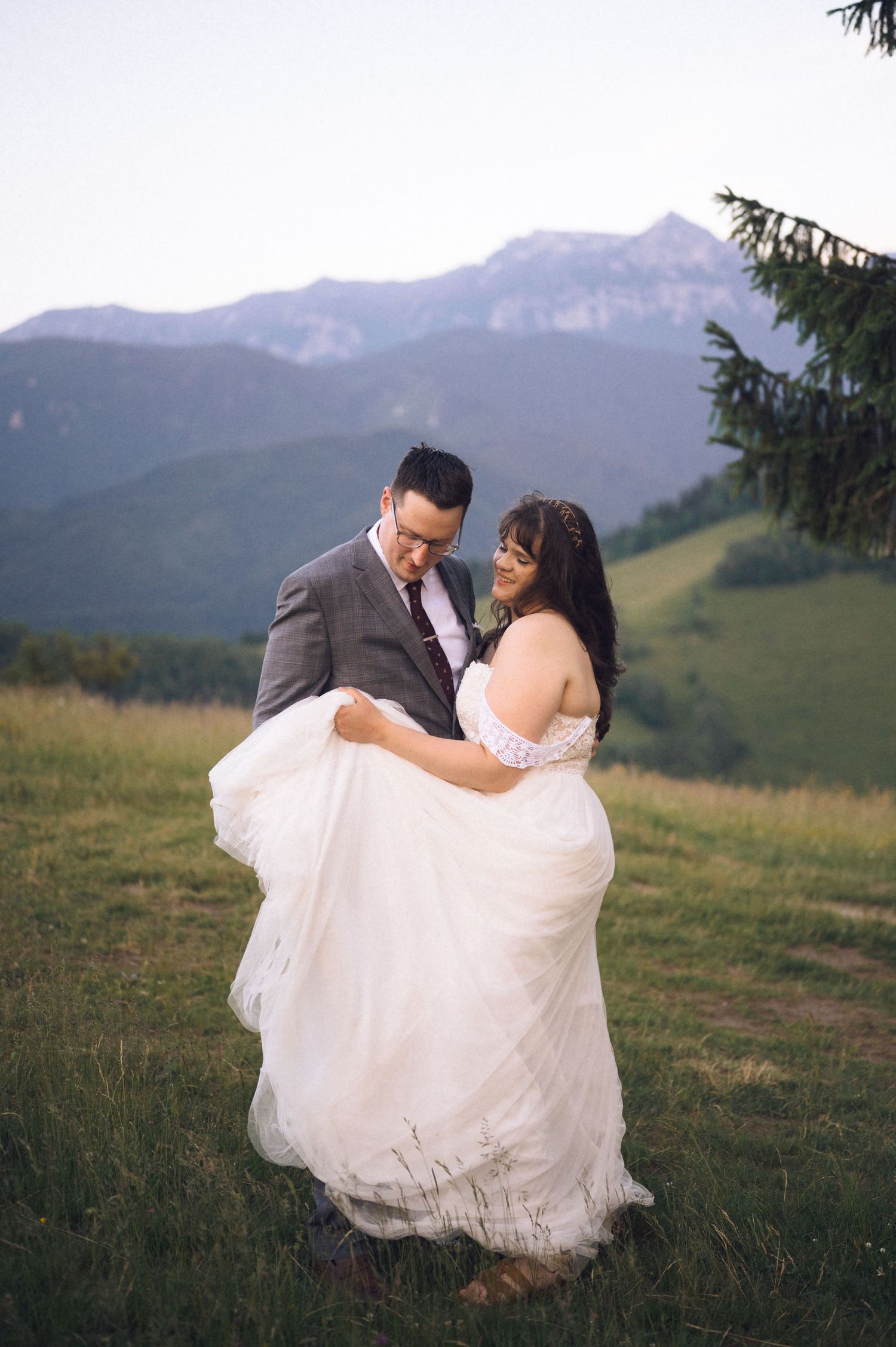 Groom lifts bride in white off-shoulder gown on mountain meadow at dusk.