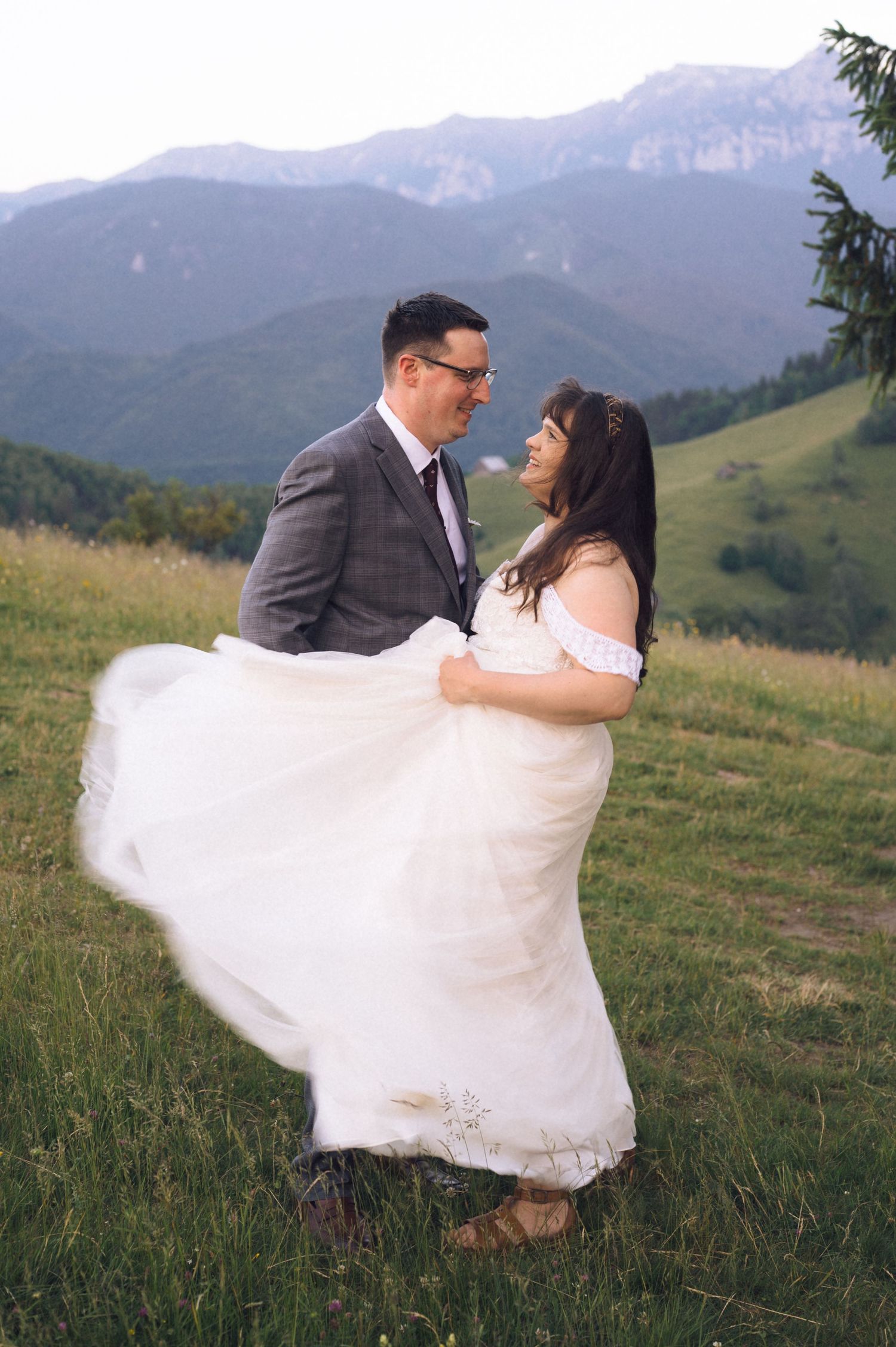 Couple in wedding attire dancing on a green hillside with mountains in the background, bride's white gown flowing in the wind.