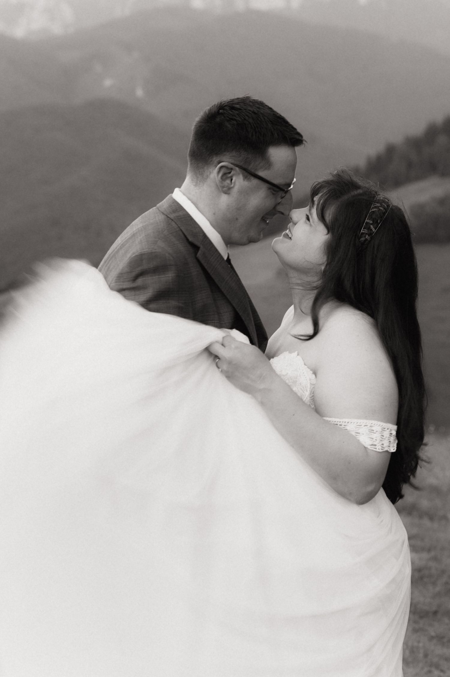 Black and white photo of a couple in wedding attire sharing a romantic moment with mountain scenery behind them.