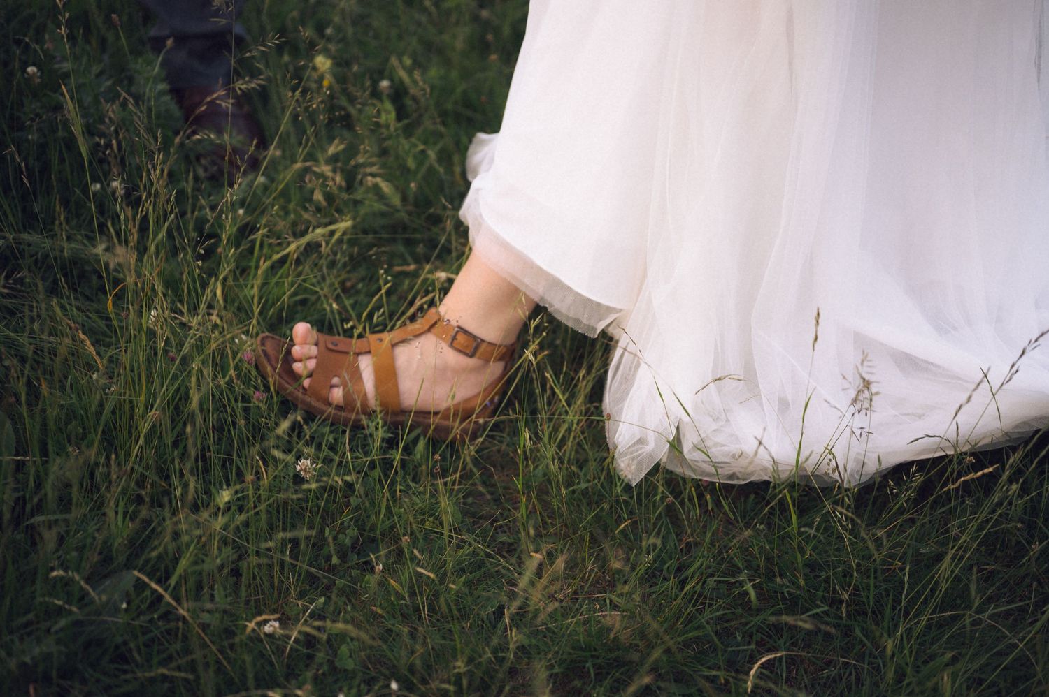 Bride wearing a flowing white wedding dress and brown leather sandals standing on lush green grass.
