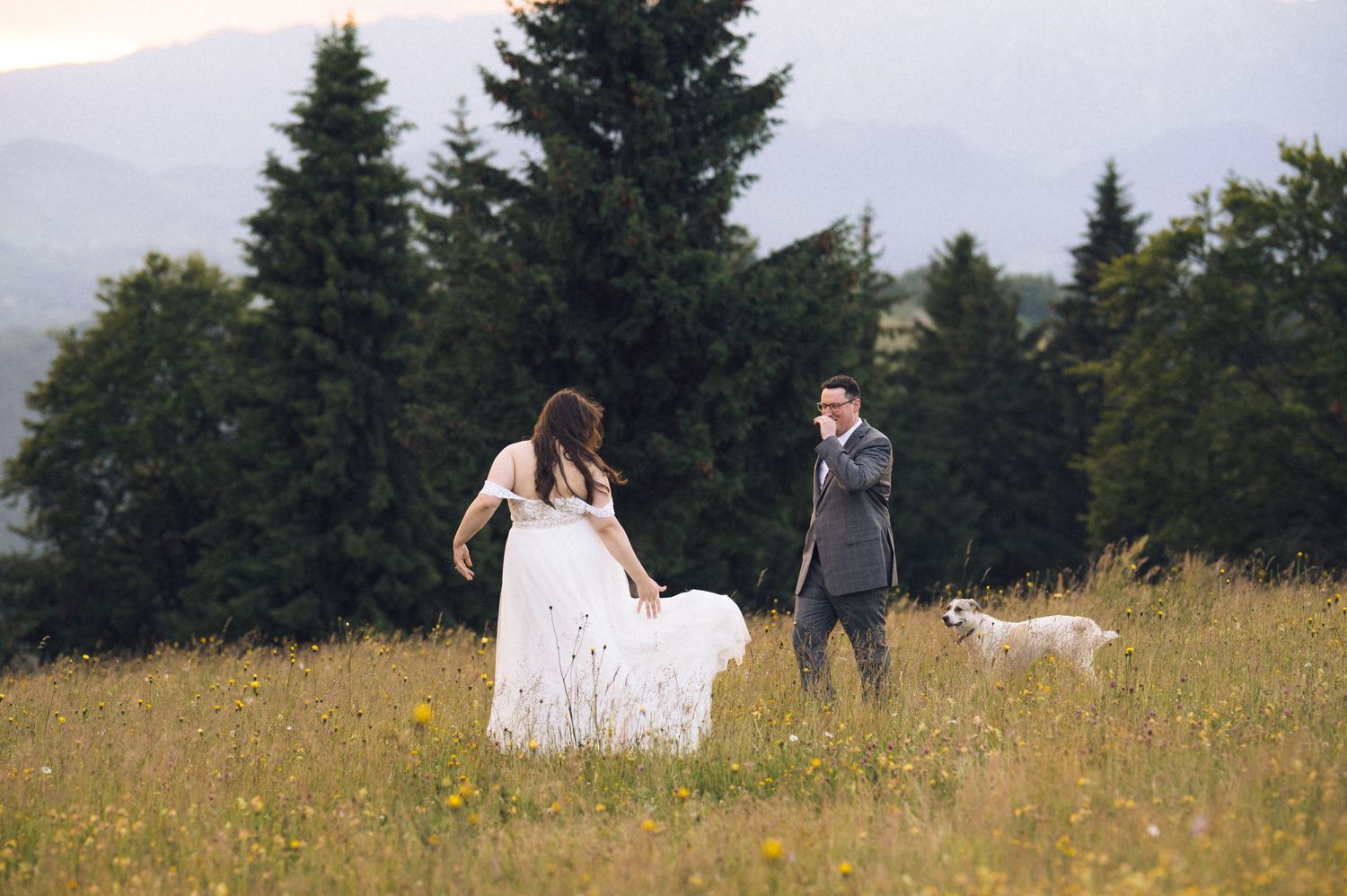 Bride in white dress and groom in gray suit walk through a wildflower meadow with a white dog and pine trees.