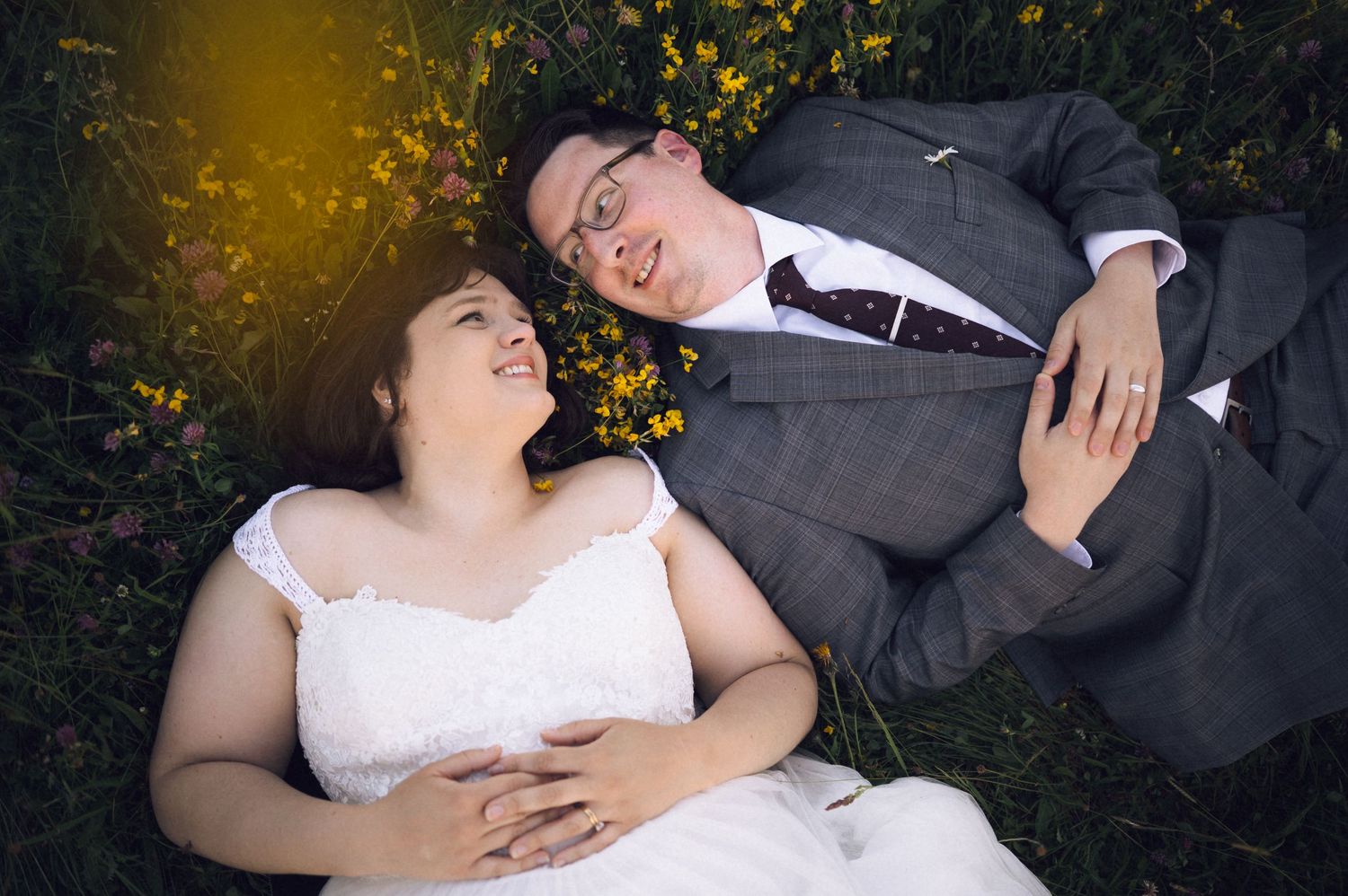 A bride and groom lying in grass surrounded by yellow flowers, smiling at each other on their wedding day.
