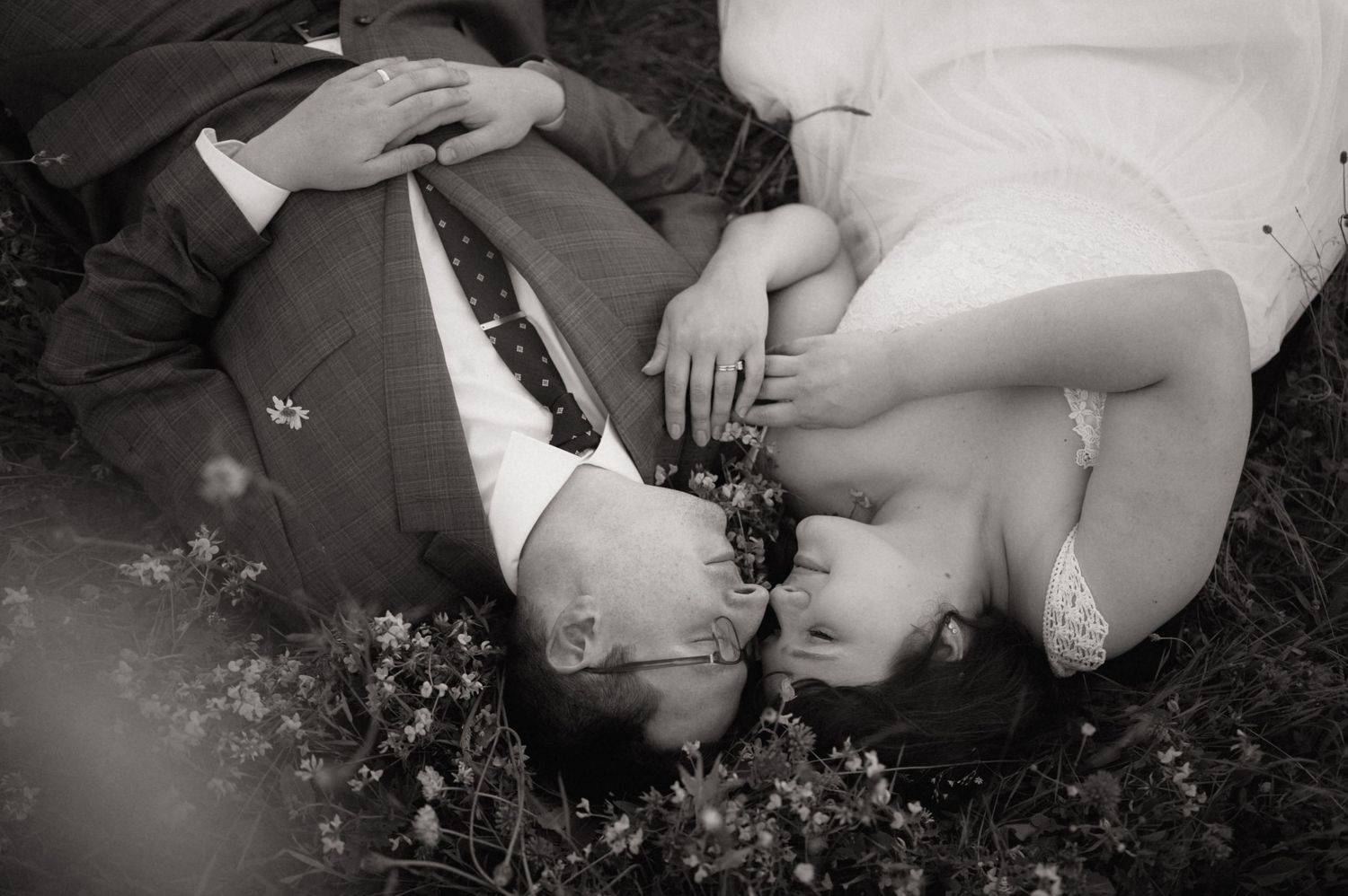 Black and white photo of a couple lying in grass, sharing a kiss surrounded by wildflowers on their wedding day.
