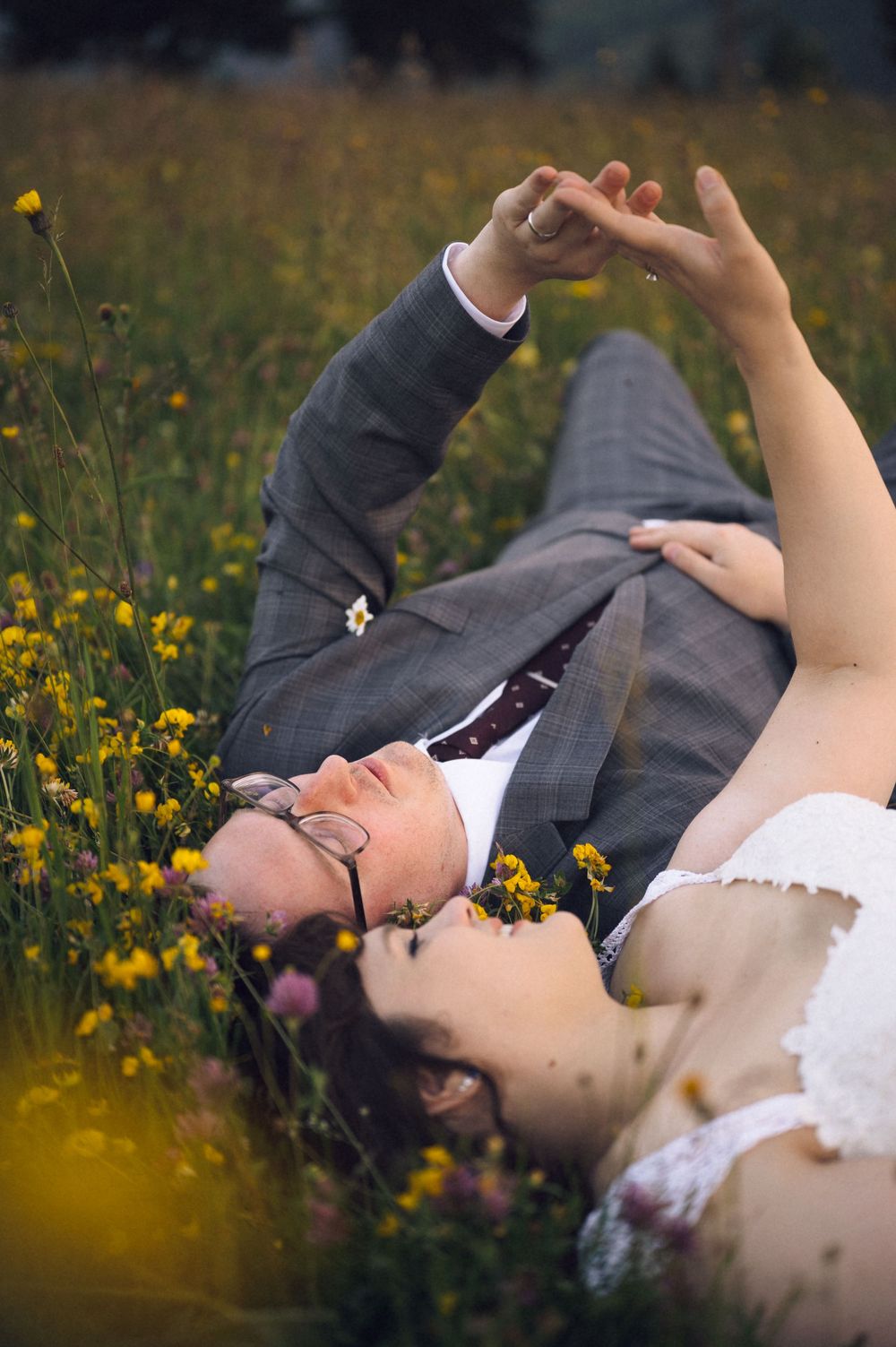 Bride and groom lying in a wildflower meadow, holding hands and gazing upward on their wedding day.