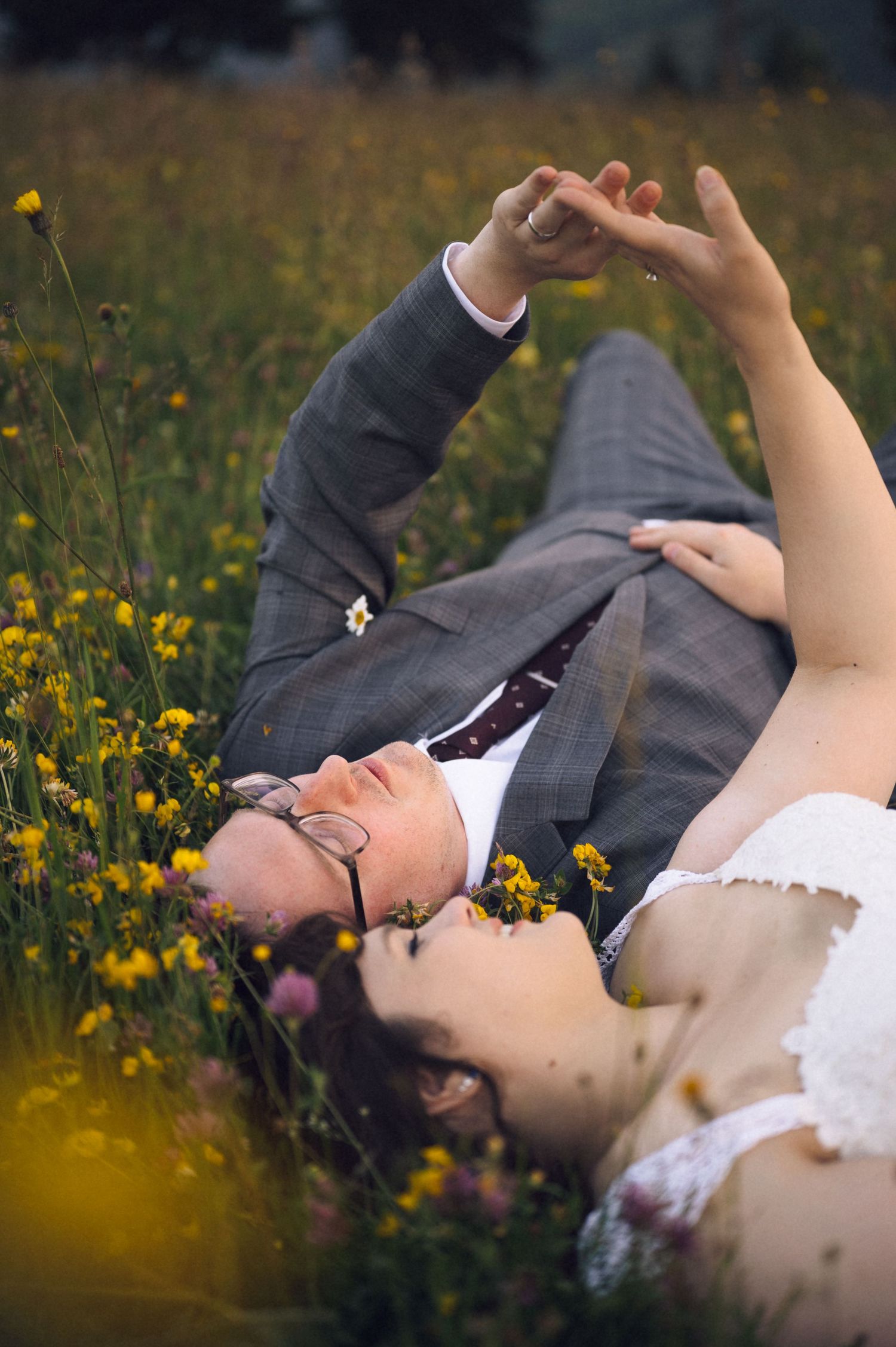 Bride and groom lying in a wildflower meadow, holding hands and gazing upward on their wedding day.
